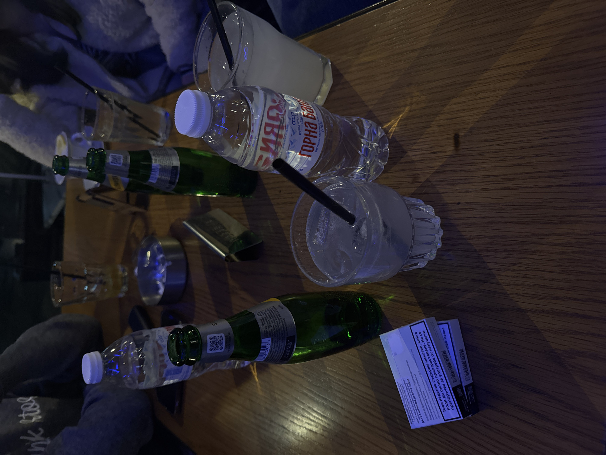 On a wooden table are two bottles of sparkling wine, a bottle of GORNA BANIA mineral water, two glasses with straws, one with a clear liquid and ice, another with a milky-white liquid, a metal object, and a pack of cigarettes with a Bulgarian inscription. The scene is likely in a bar or restaurant. The location is unspecified. The mineral water brand "GORNA BANIA" is a clue that the location is somewhere in Bulgaria, but not necessarily the city of Goce Delcev.