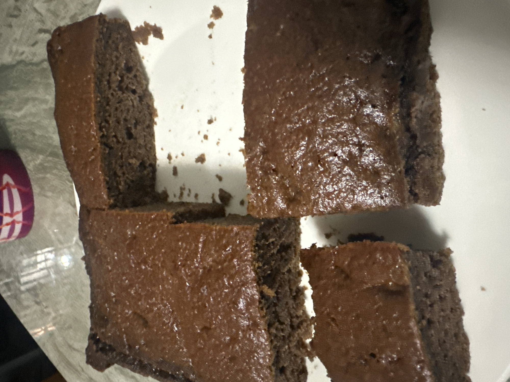 The image shows four pieces of chocolate cake on a white plate. The cake appears to have a glossy, smooth frosting. Crumbs are scattered around the cake pieces on the plate. A small portion of a container with a red and white pattern is visible on the left side of the image. The background is a light gray, textured surface. The location, Bac Nha Trang Ward, Vietnam, is not discernable from the image content. There is no information about people, actions, or interactions present in the photograph.