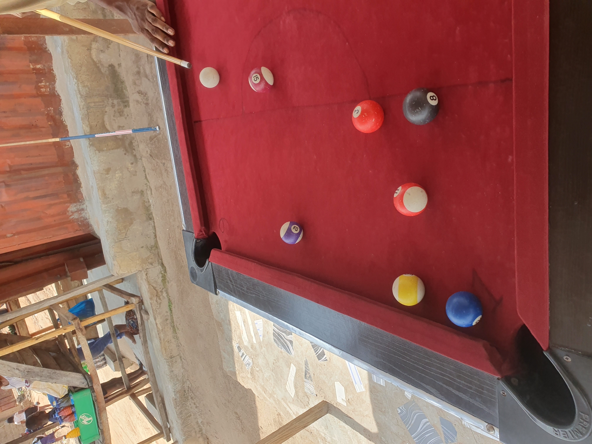 The image depicts an outdoor billiard table set up on a sandy surface near a concrete structure. Several billiard balls are scattered on the red felt surface of the table, including a white cue ball, a purple ball with the number 12, a pink ball with the number 15, a red ball with the number 3, a yellow and white striped ball, a blue ball, and a black ball with the number 8. A person's hand is visible on the right side of the image, holding a pool cue aimed at the white cue ball. In the background, a makeshift wooden structure with a red corrugated metal roof is visible. The environment appears to be a public space, possibly a market or recreational area, given the presence of people and the general setting. The ground is sandy and uneven, with some decorative tiles visible in the lower left quadrant. The scene is illuminated by natural daylight, casting shadows on the ground. The presence of a green plastic crate containing bottles of drinks on the wooden structure suggests a commercial or vendor setting. The overall impression is that of an informal outdoor recreational space in Lokoja, Nigeria.