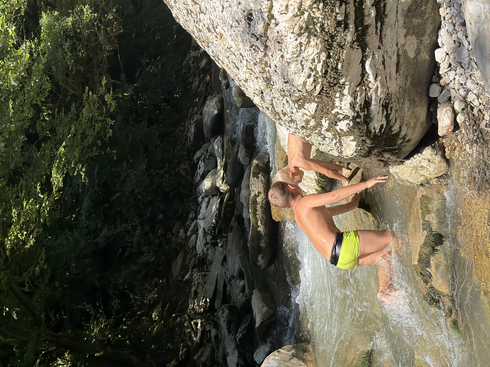 The image depicts two young boys in a shallow, rocky river in Khostinskiy, Russia. The boy in the foreground, on the left, is wearing neon green swim trunks and is kneeling in the water, facing away from the camera. His arms are extended forward, as if reaching for something. The second boy, to the right and further back, is partially obscured by a large, white, moss-covered boulder. He appears to be leaning forward, also facing away from the camera. The riverbed is composed of numerous smooth, grey and brown stones, some of which are covered in green moss. The water is clear and appears to be flowing rapidly, creating ripples and splashes. Lush green foliage and trees overhang the riverbanks, casting shadows in some areas. Sunlight illuminates the water and the upper portions of the trees, creating a dappled lighting effect. The scene is set in a natural, outdoor environment, likely a canyon or gorge, given the rocky terrain and dense vegetation.
