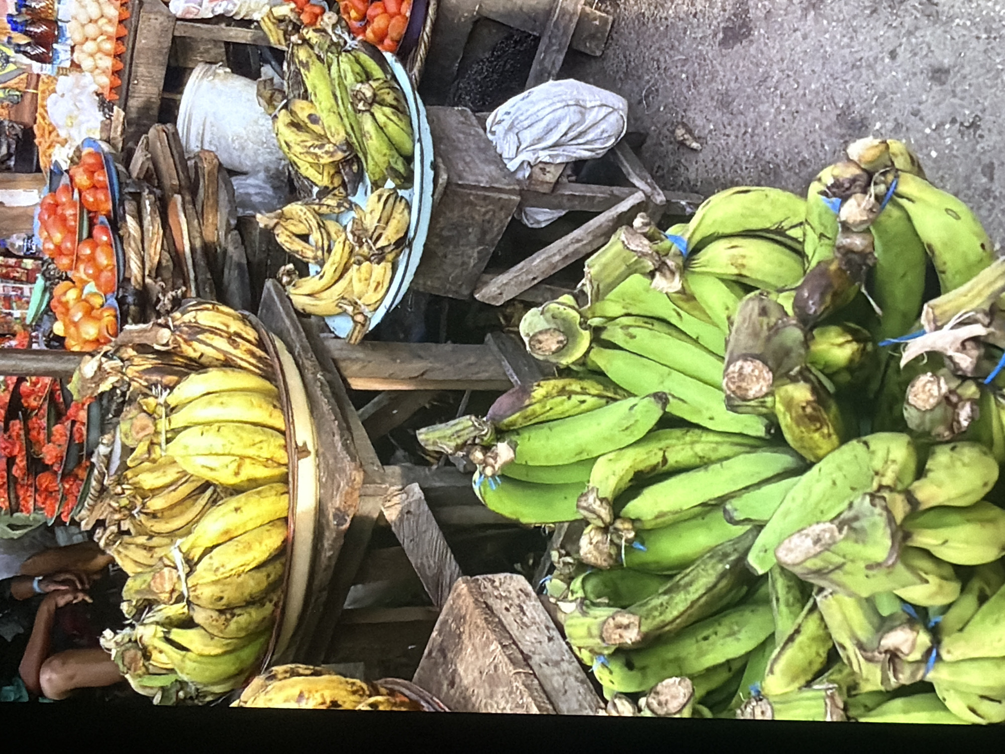 The image depicts a market stall in Bukuru, Nigeria, showcasing a variety of produce. In the foreground, numerous bunches of green plantains are piled on the ground, some secured with blue ties. To the right, a white cloth bag is hanging from a wooden structure. Behind the plantains, on a slightly elevated wooden surface, are more bananas, some yellowing, arranged in a light blue bowl. Further back, various red tomatoes are displayed in bowls. The background reveals more market stalls with assorted goods and indistinct figures of people. The ground appears to be a rough, earthy surface.
