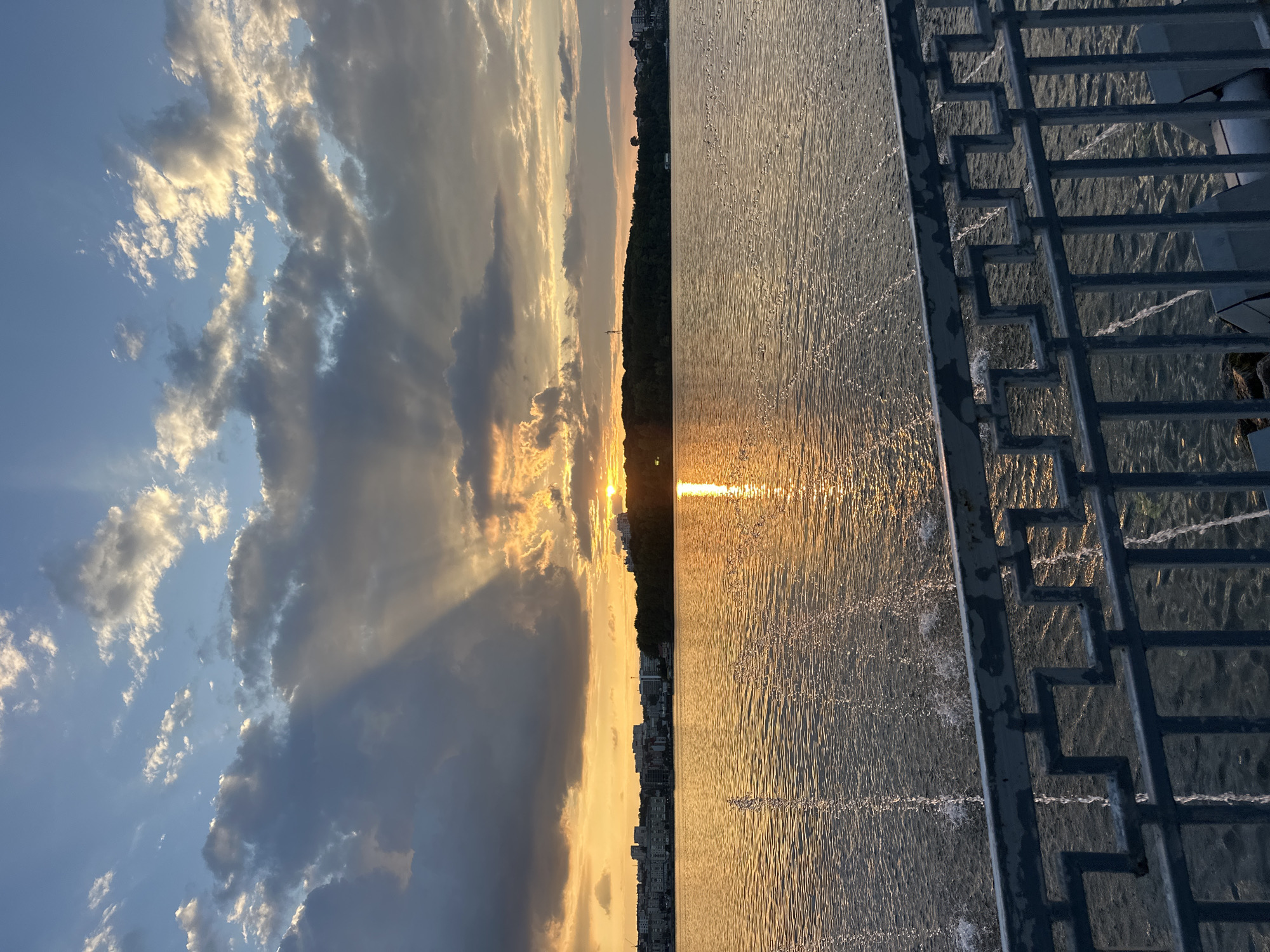 The image depicts a sunset over Ternopil Pond in Ternopil, Ukraine. The sun, a bright orb, is low on the horizon, casting golden rays through scattered clouds. The water of the pond reflects the warm hues of the sunset. In the foreground, a metal railing with a geometric pattern runs across the bottom of the frame. Water jets from fountains are visible in the pond. Along the far bank of the pond, a line of buildings and trees forms the city skyline. The sky transitions from a bright blue near the top to shades of orange and yellow near the horizon, with dark clouds casting shadows and creating dramatic light effects. There are no people visible in the image.