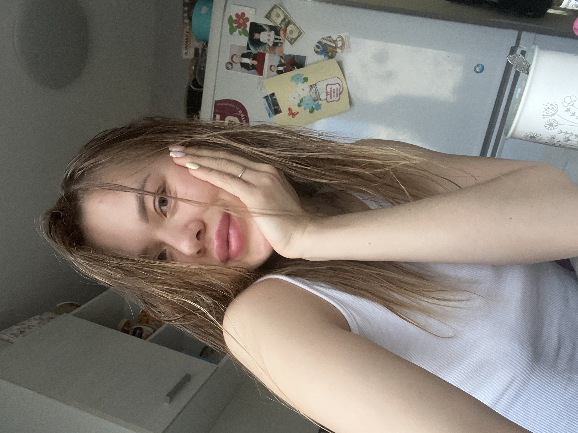 A young woman with light brown hair and a white tank top is posing for a selfie indoors. She is holding her face with her hand, displaying a ring on her finger. Behind her is a white refrigerator decorated with magnets and photos. In the background, there is a white cabinet. The location is likely an apartment in Chaikovsky, Russia.