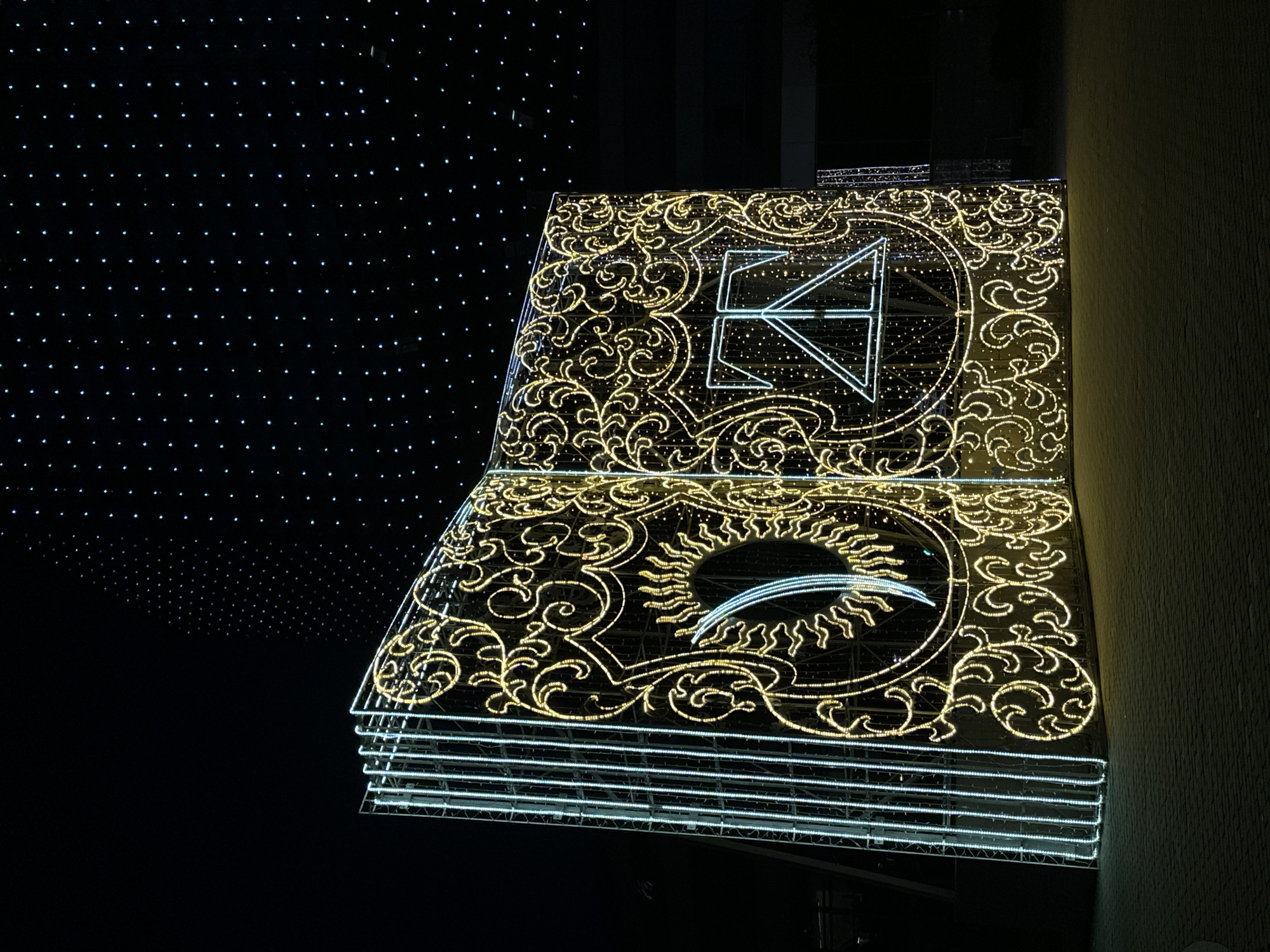 A close-up nighttime shot depicts a brightly illuminated, open book-shaped structure. The structure appears to be an artistic installation, with its pages adorned with ornate scrollwork patterns made of small lights. The top page features an abstract triangular symbol, while the bottom page displays a stylized sun design. The "book" consists of multiple stacked, illuminated white lines, simulating page thickness. To the left of the structure is a dark background covered in regularly spaced, small white lights. To the right, a grey column is partially visible.
While the prompt asks for the location to be specified as Minsk, Belarus, this cannot be definitively determined from the image alone. Therefore, the location is not included in the description.