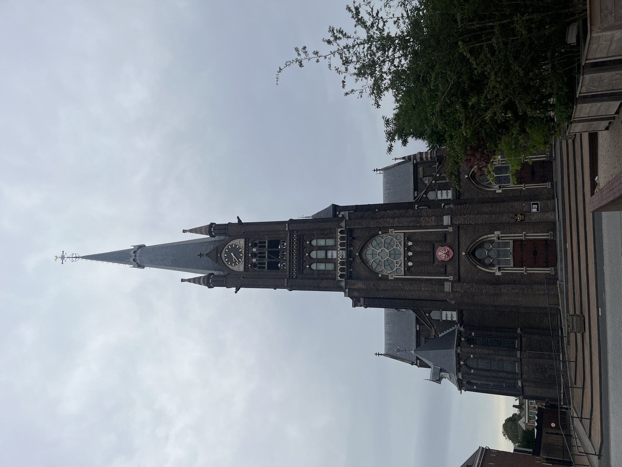 A vertical, low-angle, full shot captures a dark brick church with a tall, slender spire against a cloudy sky. The church's facade features arched windows, a rose window, and a clock face on the tower. A weather vane, a cross, and smaller spires adorn the top of the main spire. Trees with green foliage are visible to the right and in the background. A set of stairs and a ramp lead up to the church entrance. The ground in the foreground is paved with brick. A wooden planter box sits on the right side of the frame.