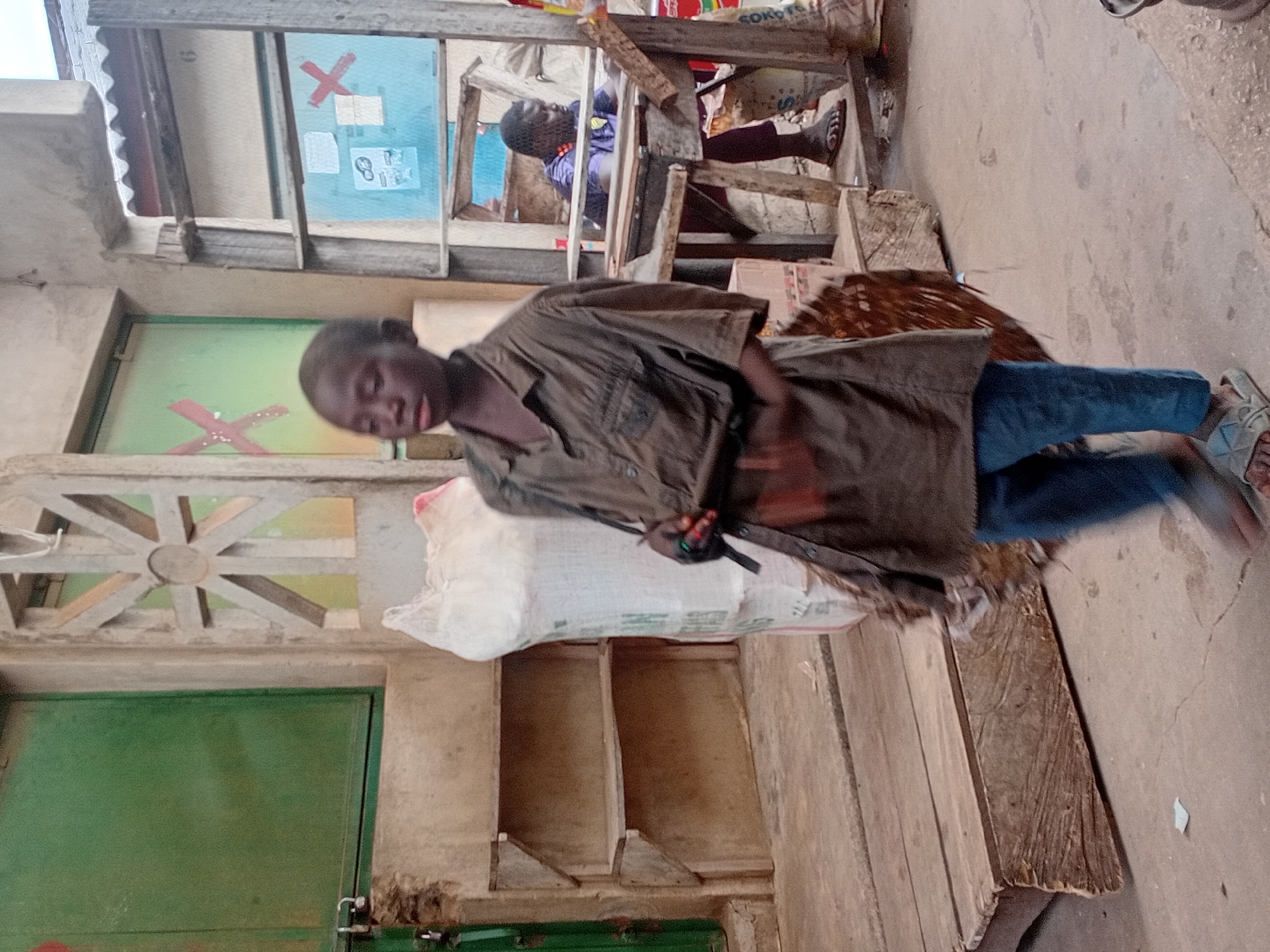 In a market setting in Maiburiji, Nigeria, a young boy stands in the foreground, facing slightly to the right. He wears a loose-fitting, dark-colored, button-up shirt and blue pants. His feet are clad in light-colored sandals. He is holding a dark object with a strap across his chest, possibly a bag or a tool. Behind him, to the left, is a large, white sack. Further back, a wooden shelf unit is visible against a textured wall. To the right of the boy, an older man sits at a wooden structure. He is wearing a purple shirt and dark pants, with sandals on his feet. He is looking down at something in front of him. The environment appears to be an open-air market with various structures, including a green door to the far left and a window with a red "X" marking on it behind the boy. The ground is concrete.