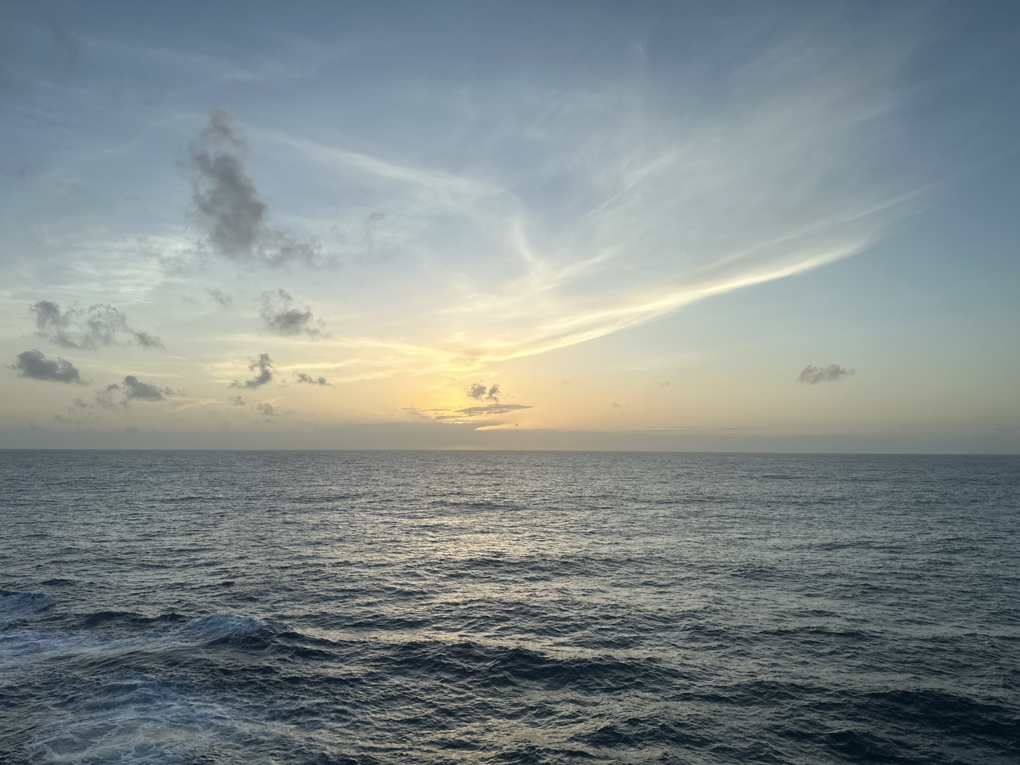 The image shows an ocean scene under a sky transitioning from blue to shades of yellow and orange at the horizon, suggesting sunrise or sunset. The ocean is characterized by small waves, with a visible texture caused by sunlight reflecting off the water's surface. The sky is partially covered with clouds, some dark and dense, others thin and wispy. The location is over open water, with no land visible. The city and state are unknown.