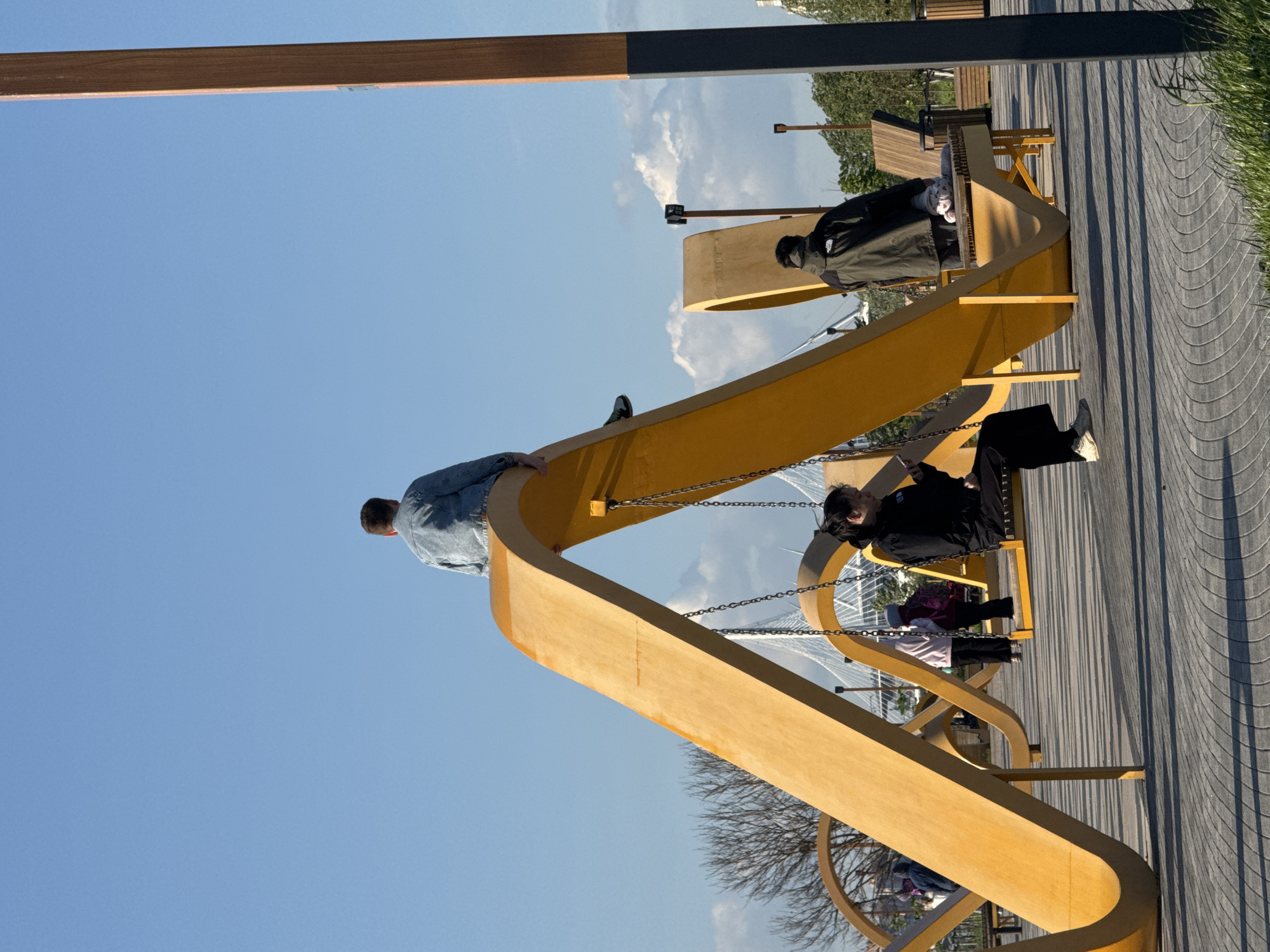 The image captures a playful, contemporary public space with people engaging with artistic swing sets. It is a sunny day, and the sky is clear blue.
**People:** The photo contains several people in varying states of interaction with the swings. A man in a denim jacket sits on the curved top of one of the swing structures. Other people are seated on the actual swings, including a person in black and others who are partially obscured.
**Subjects/Objects:** The primary subjects are the sculptural swing sets, which are a bright, golden yellow color. They are made of curved wooden elements and suspended by metal chains. There is also a wooden boardwalk-style pathway with a wavy pattern. Bare tree branches are visible in the background, along with a view of a modern bridge with a cable-stayed design.
**Actions/Interactions:** People are sitting on the swings and on top of the swing structure.
**Environment:** The scene takes place in an outdoor urban setting, likely a park or promenade area. The presence of the modern bridge indicates a contemporary urban design.
**Location:** The cable-stayed bridge in the background, with its distinctive A-shaped pylons, is likely the cable-stayed bridge ZSD, near the Lakhta Center in Saint Petersburg, Russia. The surrounding environment appears to be a public recreational space near the Gulf of Finland.