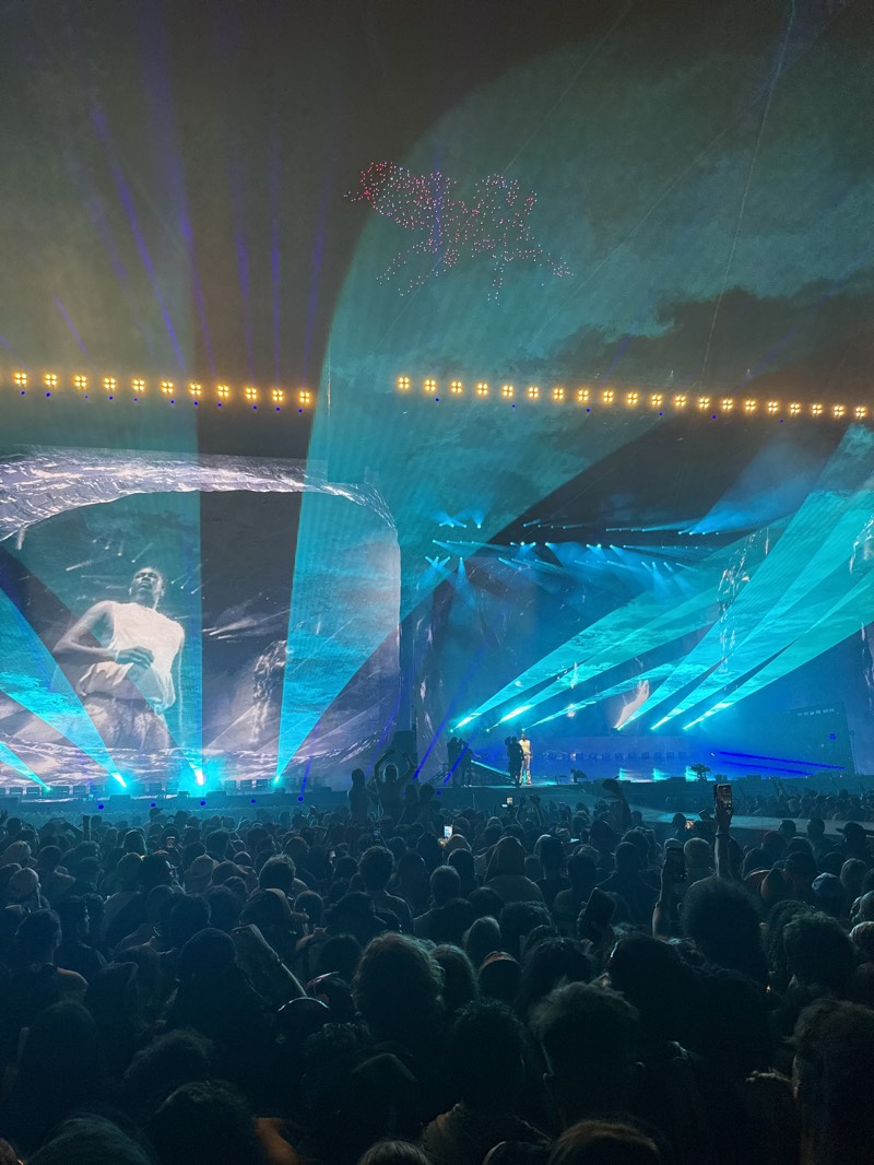 The image depicts a large crowd of people at a concert, likely the Coachella Valley Music and Arts Festival, located in Indio, California, United States. The crowd faces a brightly lit stage with a large screen displaying a black and white image of a person. On the stage, figures appear performing. Above the stage, there are stage lights and a drone show forming the image of a Pegasus. The environment is dark except for the stage lighting and screens. Many in the crowd are holding up phones.