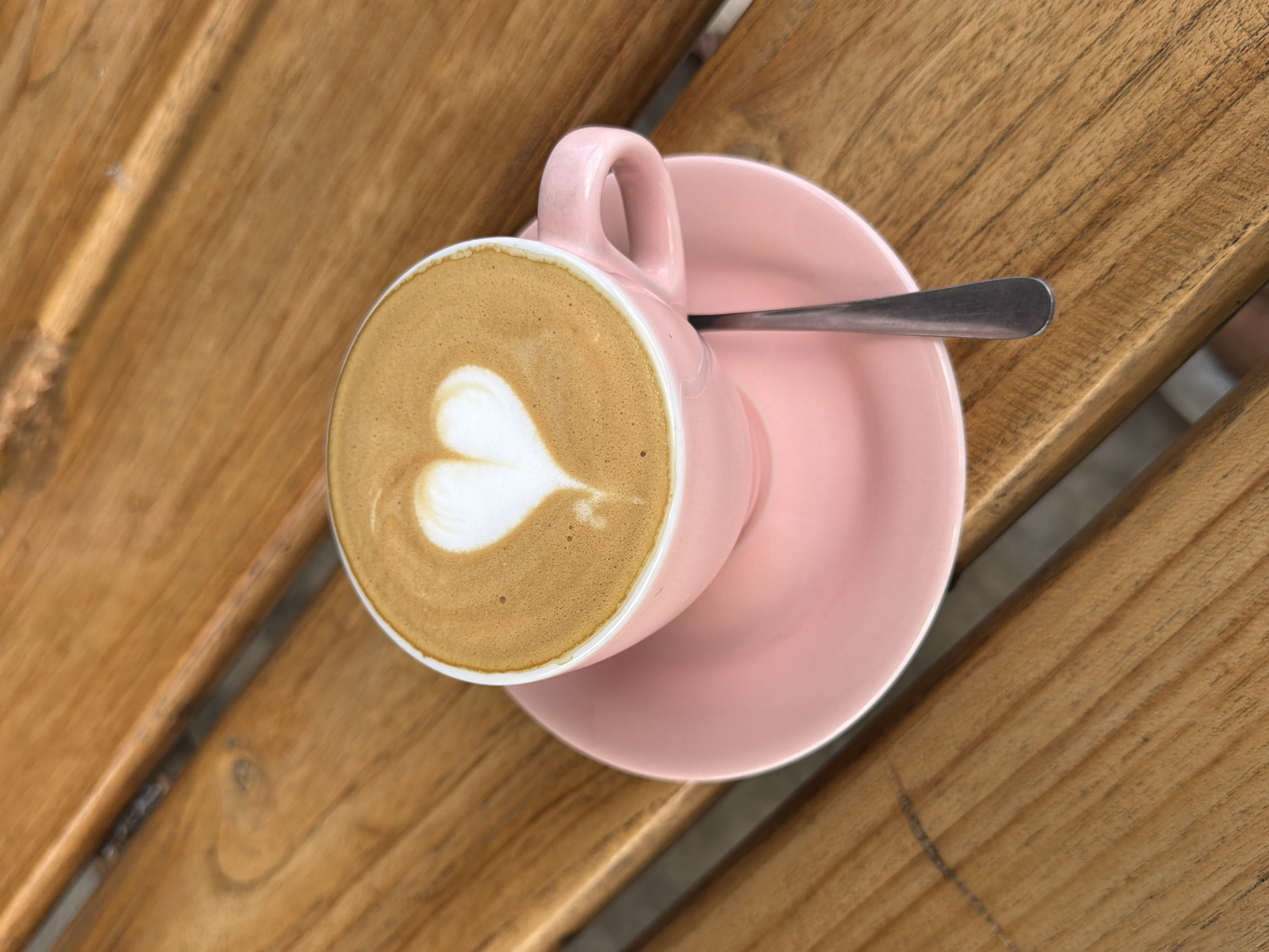 A pink cup of coffee with latte art in the shape of a heart sits on a pink saucer, which is placed on a wooden table. A silver spoon rests on the saucer. The image appears to be taken at a cafe in Mengwi, Indonesia.