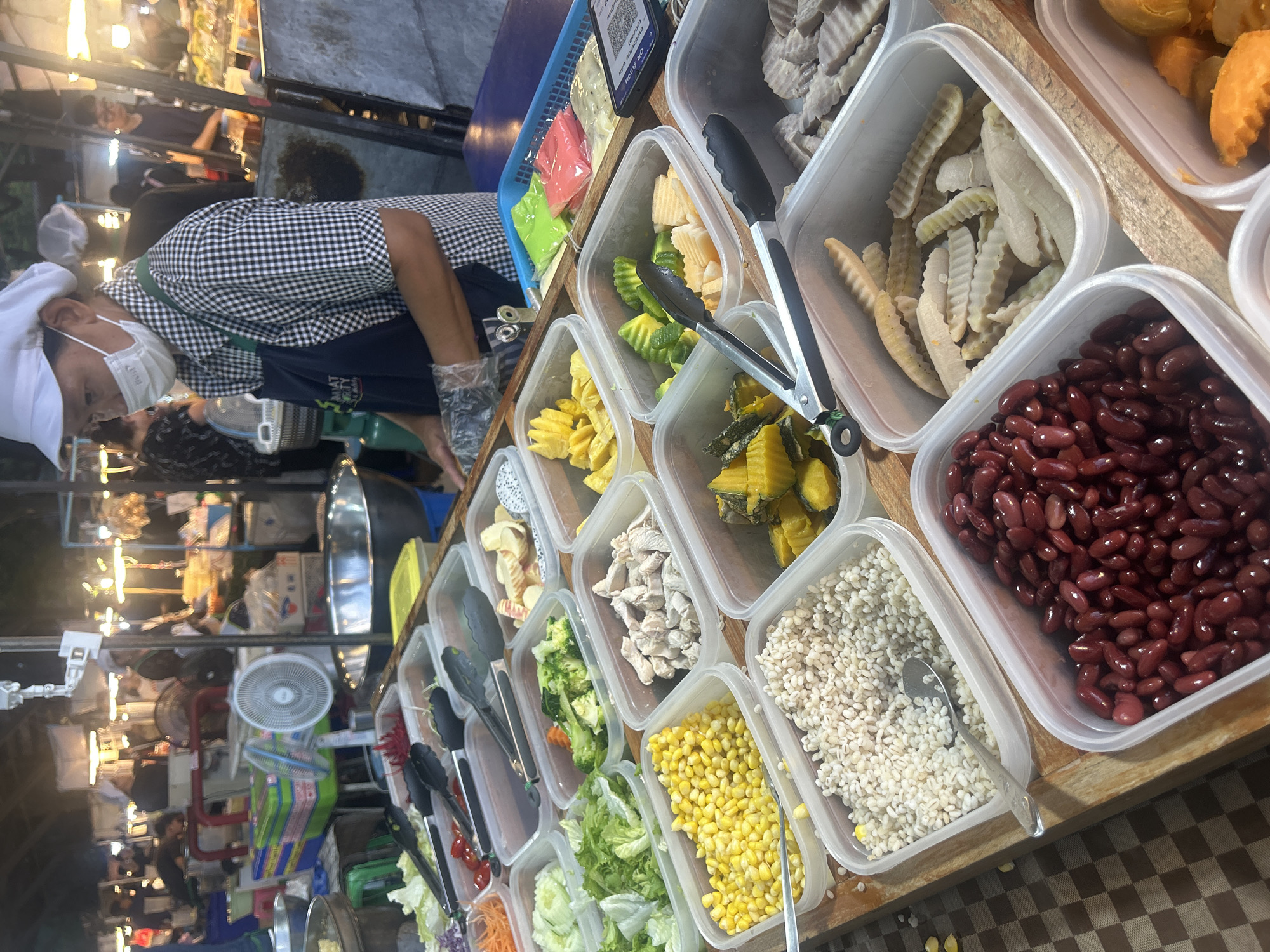 This is a medium shot of a food stall at a street market in Tha Khlong Town Municipality, Thailand. The stall features an array of food items displayed in clear plastic containers. In the foreground, there are containers of red kidney beans, what appears to be barley or a similar grain, and corn kernels. Behind these, other containers hold chopped chicken, broccoli florets, sliced cucumbers, shredded carrots, lettuce, and what might be pumpkin. Further back, there are containers with cut fruits like pineapple and dragon fruit, and some other unidentifiable processed food items. A person wearing a white cap and a checkered shirt with a dark apron is visible in the background, appearing to be serving food or preparing an order. They are also wearing a face mask and gloves. The stall is brightly lit by overhead lights, typical of a night market. The overall environment suggests a busy, open-air food market.