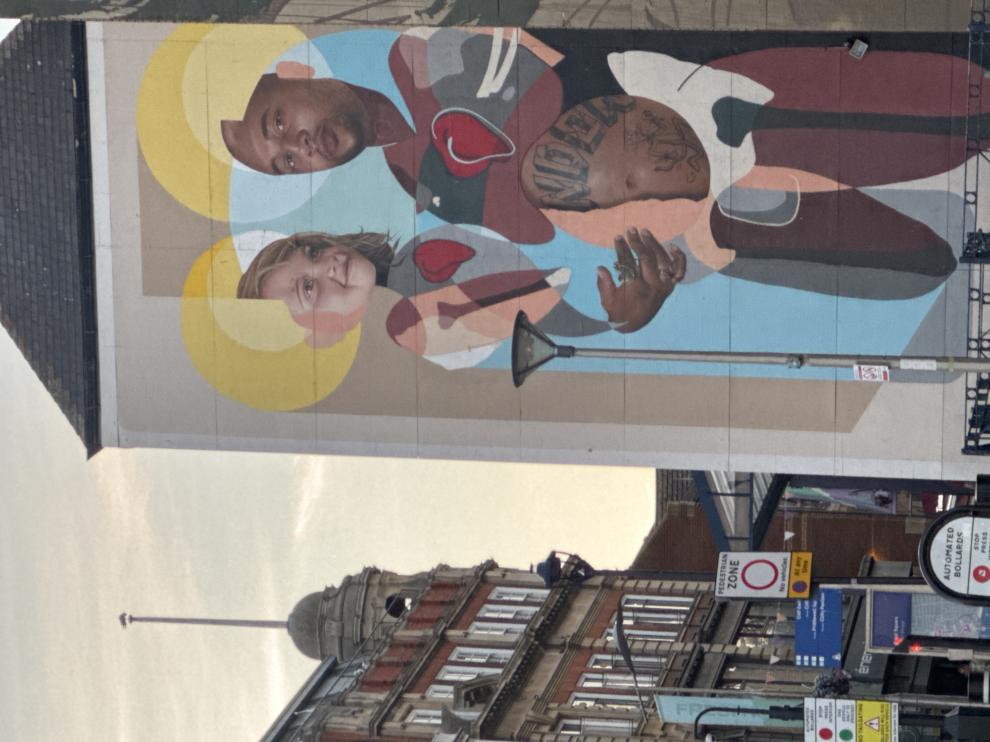 The photograph captures a vibrant mural on the side of a building in Southend-on-Sea, United Kingdom. The mural depicts three figures: a young girl with blonde hair and a smiling expression, a man with dark skin and short hair, and a partially visible individual beneath the man's arms, whose face is covered in tattoos. The figures are stylized with abstract shapes and pastel colors, with red hearts prominently placed on the chests of the girl and the man. Both the man and the girl have yellow circular halos above their heads. The man is wearing a dark garment with grey and red accents.
To the left of the mural, an old brick building with multiple stories and numerous windows is visible, featuring architectural details like arched windows and decorative cornices. A flagpole stands atop a dome-like structure on this building. Several street signs are also present, including a "PEDESTRIAN ZONE" sign with a red circle and line indicating no vehicles, and other signs providing directions to various locations and mentioning "automated bollards." A streetlamp with a black shade is mounted on a pole to the left of the mural. The sky in the background is overcast with soft, diffused light, suggesting it might be late afternoon or early morning. The overall scene is a street view combining urban architecture with contemporary street art.
