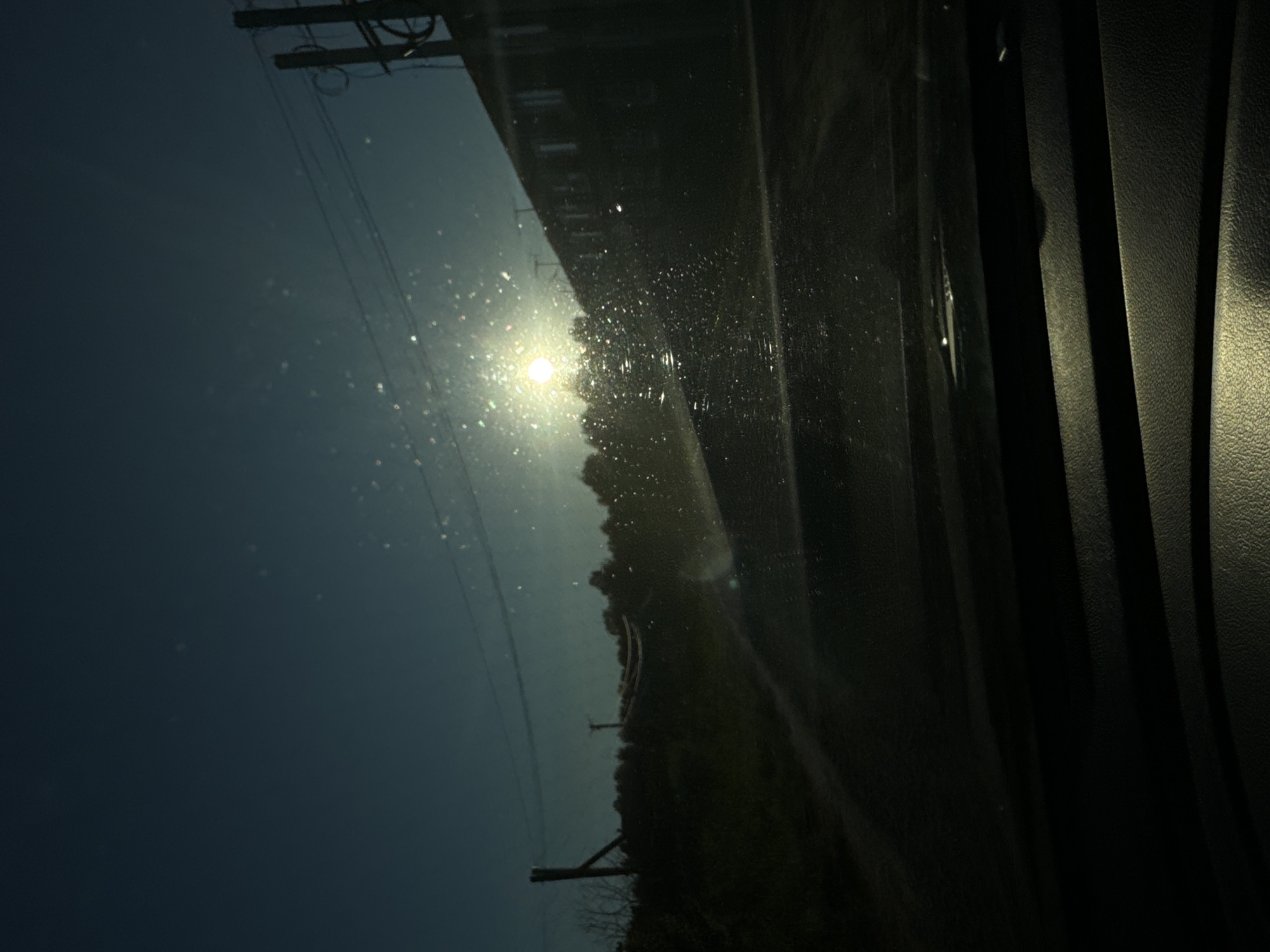 The image appears to be taken from inside a vehicle, looking out through the windshield. The sun is low in the sky, casting a bright glare and creating numerous light refractions and dust particles on the glass. In the background, a dark, tree-lined hill is visible, with a building or series of buildings silhouetted against the sky on the right. Power lines stretch across the sky, supported by poles. The foreground of the image is dominated by the dark interior of the vehicle, possibly the dashboard. The overall scene suggests a rural or semi-rural setting in Russia, possibly Ponomaryevo, during either sunrise or sunset, with the sun's low angle creating a dramatic and somewhat hazy effect.