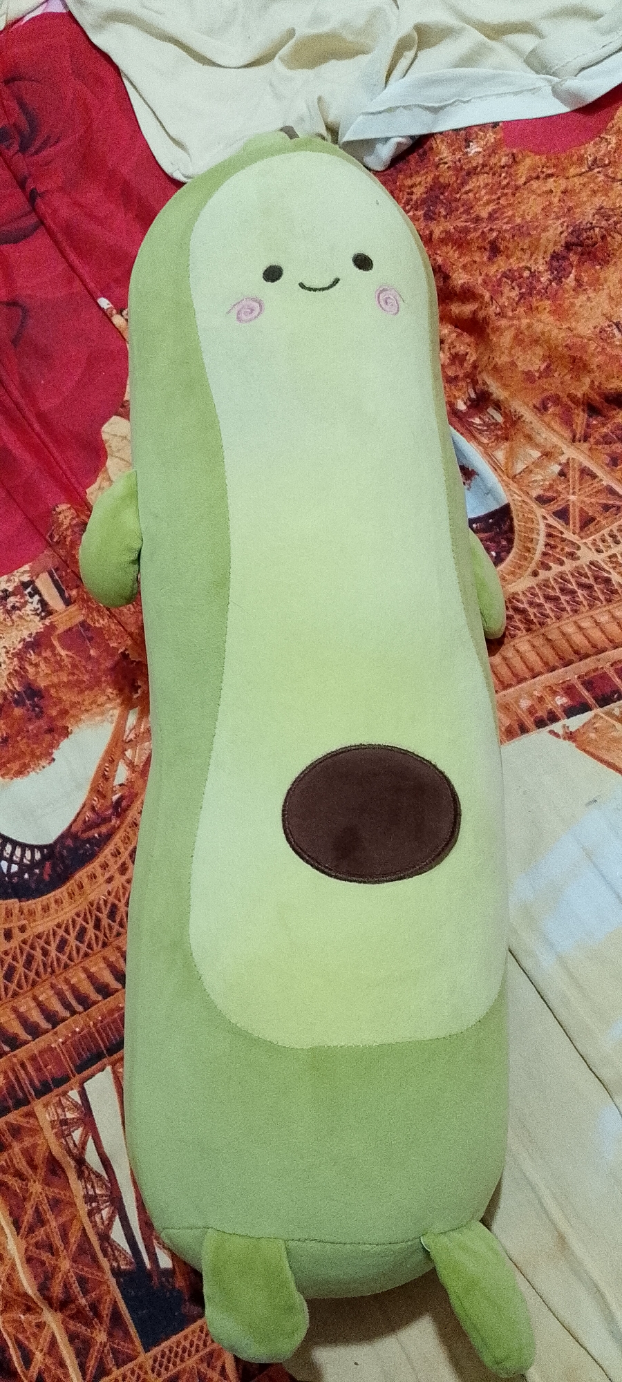 A plush avocado toy lies on a patterned bedspread. The toy is light green with a darker oval shape representing the pit in the center. It has two small arm-like protrusions on the sides, two small foot-like protrusions at the bottom, and a simple smiley face with two dots for eyes and a curved line for a mouth, along with two pink circular blush marks on its upper portion. The bedspread has a repeating pattern that appears to be a stylized Eiffel Tower motif in brown and orange hues, with some lighter beige fabric also visible. The image is taken from a top-down perspective. The location is described as Singkawang, Indonesia, though no visual elements in the image confirm this specific geographical location.