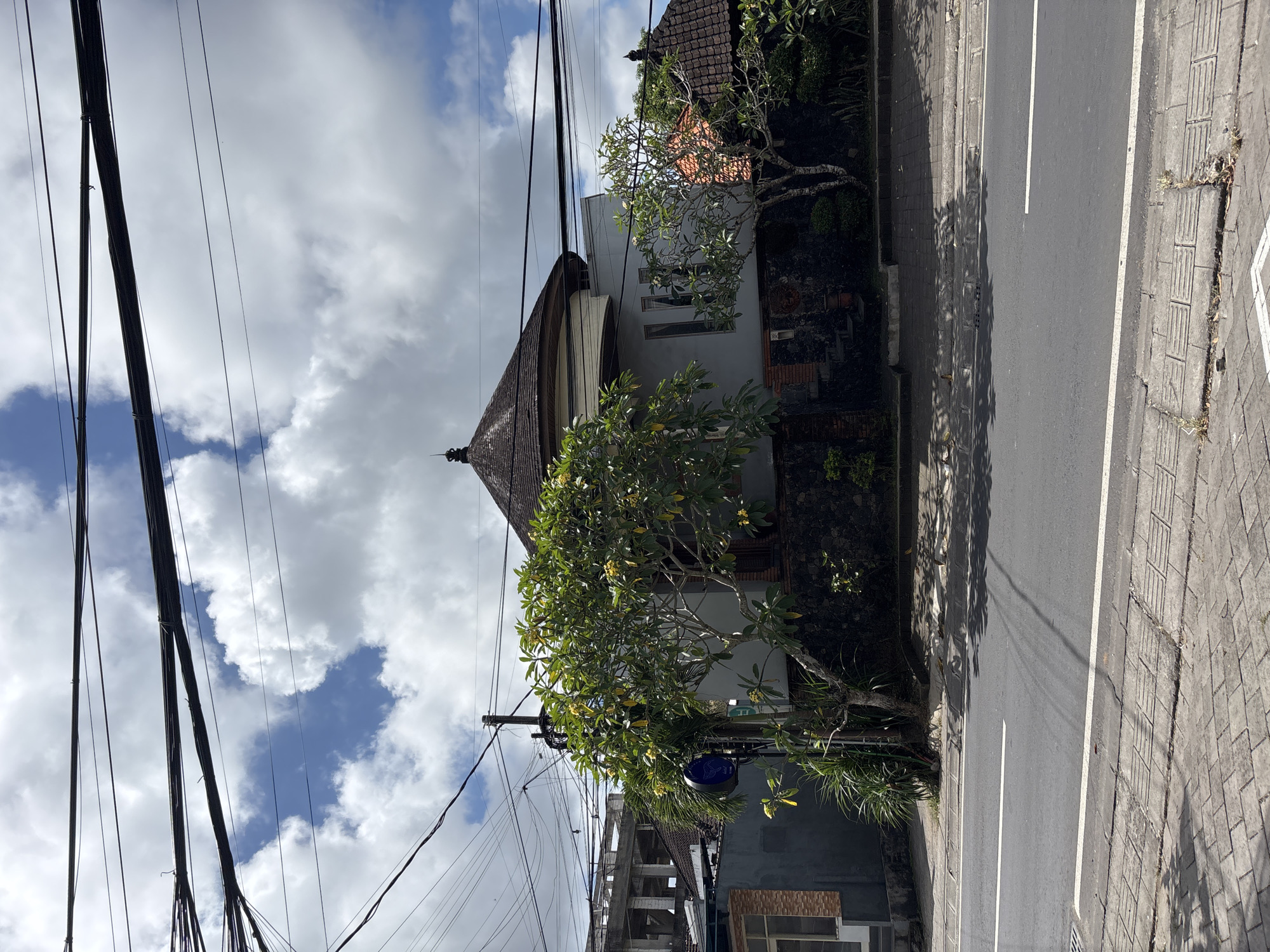 The image shows a street scene in Kuta Selatan, Indonesia. A single-lane road with a white dividing line runs along the right side of the frame. A sidewalk made of interlocking concrete bricks is adjacent to the road. A Balinese-style building with a dark tiled roof and white walls stands on the left side of the road. A frangipani tree with yellow flowers is planted in front of the building. Power lines are visible against a cloudy sky. The overall scene appears to be a typical street view in a residential or commercial area of Kuta Selatan.