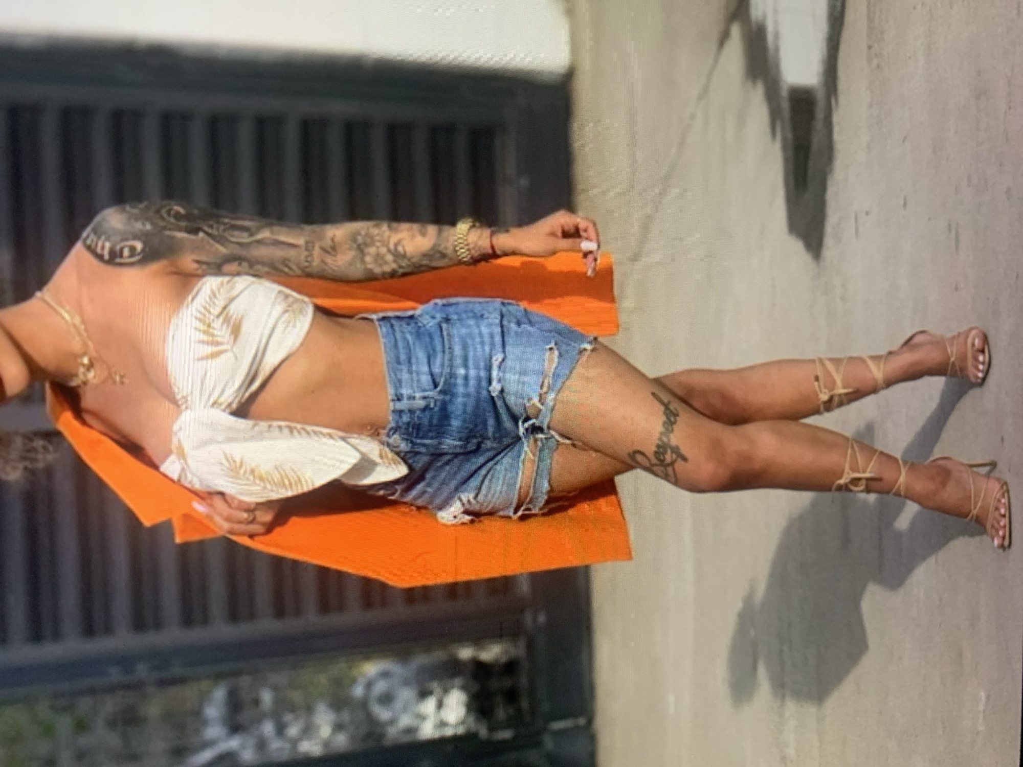 In an outdoor urban environment with a blurred dark background, a woman stands facing forward. She is wearing an orange blazer draped over her shoulders, a white and gold patterned strapless top tied in the front, and distressed denim shorts. Her legs are adorned with tattoos, including the word "Respect" on her left thigh, and she wears tan strappy high-heeled sandals that wrap around her ankles. A gold watch is visible on her right wrist. The ground is a light gray, and a shadow of the woman is cast on it.