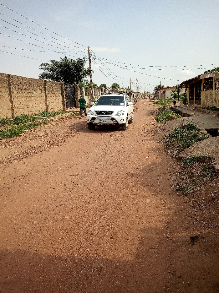 The image shows a white SUV driving on a dirt road in Ota, Nigeria (latitude: 6.691, longitude: 3.177). The road is lined with buildings on both sides, some of which have visible windows. There is a person walking in the background to the left of the car. The car has a black bumper, black wheels, and black side mirrors. The car has a license plate that reads “EPE”. The sky is overcast. There are multiple power lines running across the image. There is green vegetation on either side of the road, as well as behind the car. The ground appears dusty and brown.