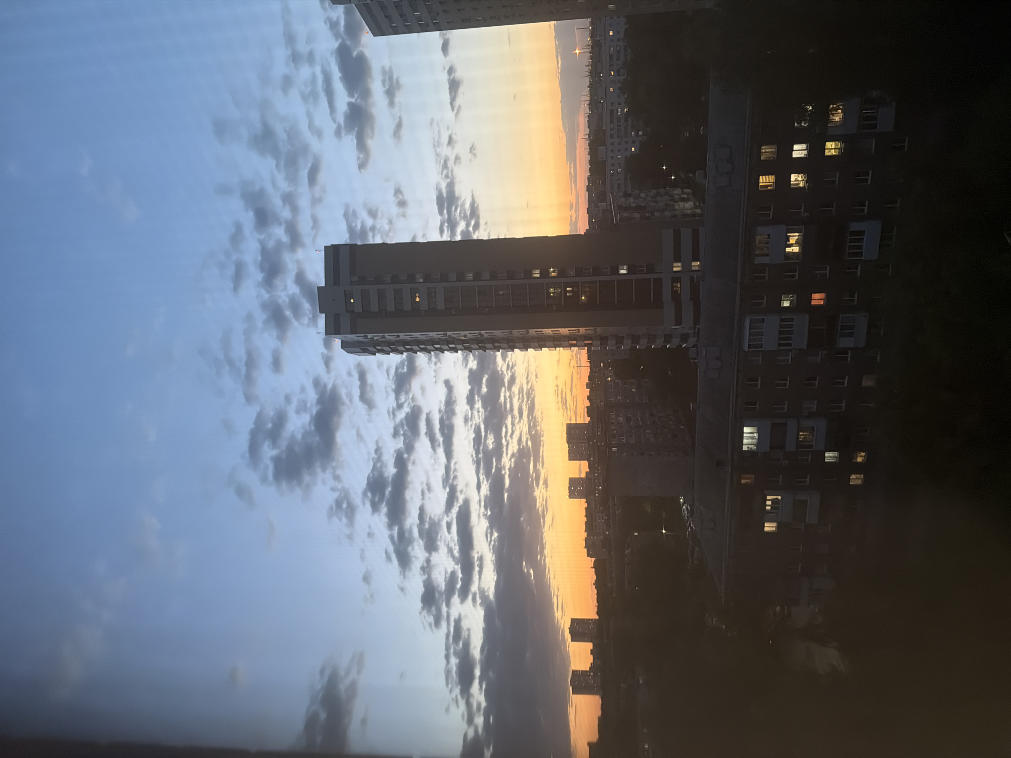 The image captures a cityscape at dusk. The sky transitions from blue to orange along the horizon, with scattered clouds. Several tall buildings dominate the scene, some with illuminated windows, suggesting residential or office occupancy. The surrounding environment is dark, indicating that it's either night or late evening. The image is taken from an elevated position, looking out over the city.