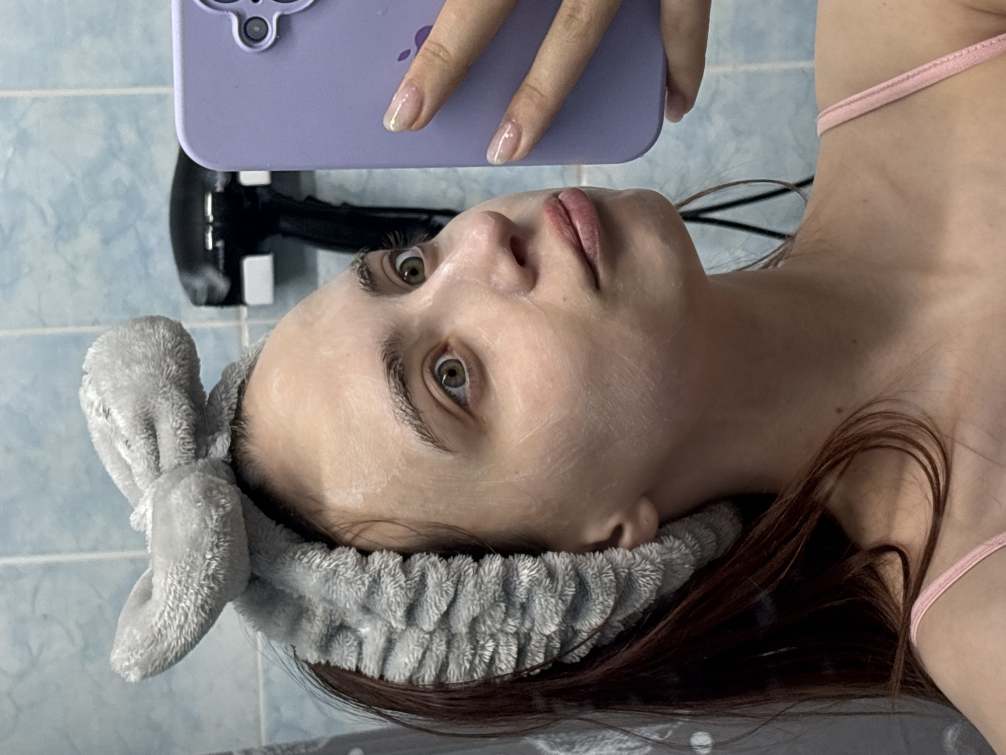 A young woman takes a selfie. She has a white cream or mask on her face and a gray headband with a bow. She is holding a purple iPhone. Her nails are manicured. The location appears to be a bathroom, with blue tiles visible in the background. A black hair dryer is also visible.