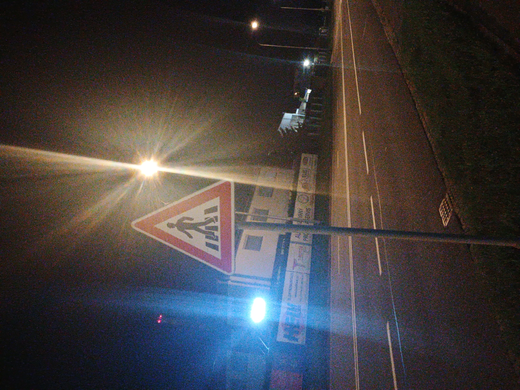 This media file captures a grainy, nighttime scene of a street or road, characterized by strong artificial light sources and dark surroundings. The image, when correctly oriented, shows a road receding into the distance from the bottom-left to the top-right.
The primary subject in the foreground (left side when oriented) is a prominent, triangular pedestrian crossing sign with a red border and a black pedestrian figure on a white background. Below and slightly to the right of this sign is a very bright blue light source, causing significant lens flare. Further down the road, several streetlights cast bright yellow-orange light, creating starburst effects and contributing to the overall illumination, which contrasts sharply with the dark sky and unlit areas.
The road itself appears to be asphalt, marked with white solid and dashed lines defining lanes or edges. To the right of the road, a darker strip of ground, possibly grass or a dirt shoulder, is visible, with a square grate or manhole cover towards the upper-right.
Behind the pedestrian crossing sign, several other signs and structures are partially visible. Some text can be deciphered on these background signs, including parts of "ELEKTROINSTALACE," and fragments like "AL," "VANY," "SERVIS," and "PRO." A circular logo or symbol, possibly an 'O' or a gear, is also discernible.
The overall impression is one of low-light conditions, typical of night photography, leading to noticeable image noise and grain. There is no specific activity depicted; the scene is static, capturing a moment on a quiet street in Kroměříž, Czechia.
