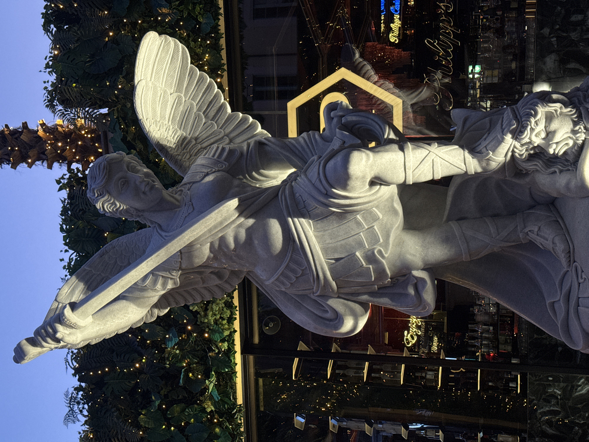 The image shows a marble sculpture of an angel slaying a demon in front of a shop window. The angel has large wings and is depicted holding a sword, while the demon is shown being defeated. The shop window reflects city lights and signs, including "Philipps" in cursive lettering. The sculpture is decorated with garland and lights. The image was taken in the city of Milan, Italy.