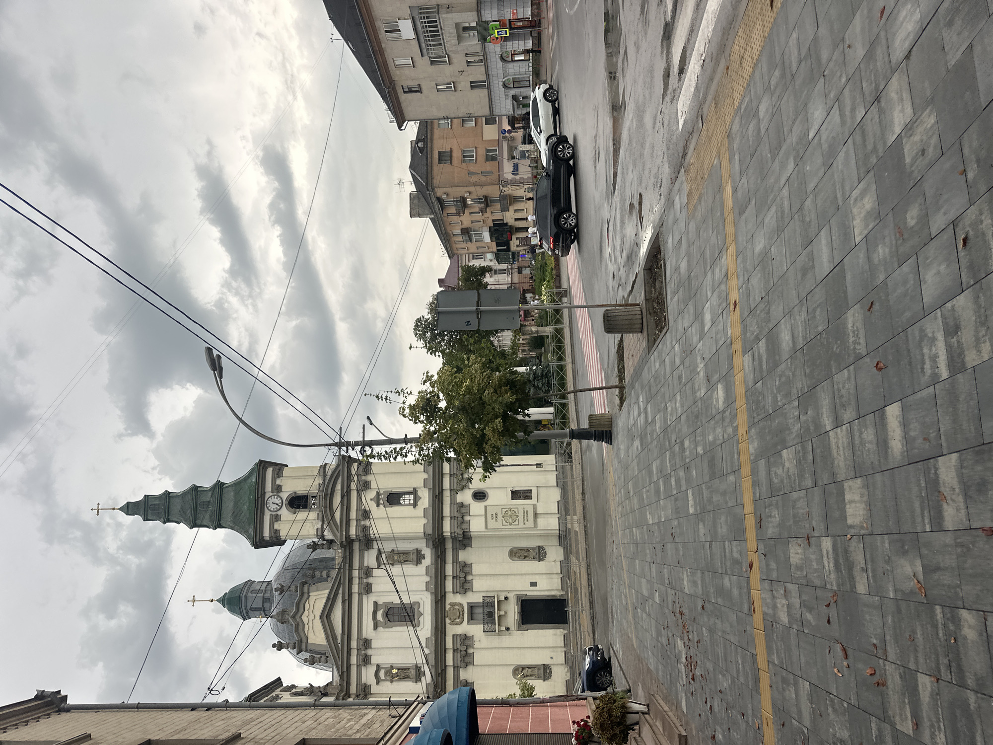 The image shows a view of the Ternopil City Hall, a historical building located in Ternopil, Ukraine. The building has a grand facade with multiple architectural elements, including domes, a clock tower, and crosses. The weather appears overcast. In the foreground, there is a pedestrian walkway with gray paving stones and fallen leaves. A street with a crosswalk is visible, and several cars are present on the road. The overall scene depicts a public space in a city, with a prominent historical building as the focal point.