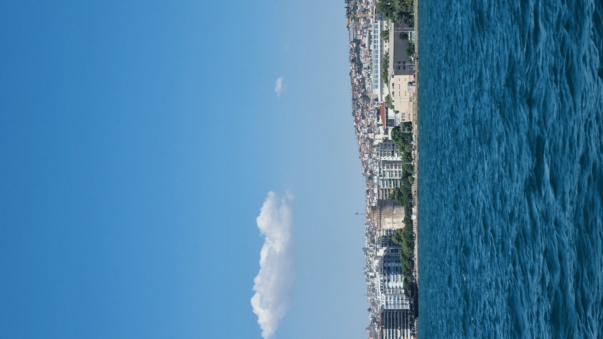 The image depicts a panoramic view of the Thessaloniki waterfront in Greece, with the White Tower prominently featured as a landmark. The scene is captured from the sea, showcasing the turquoise waters in the foreground with gentle ripples. Behind the water, a line of buildings stretches along the coastline, ranging from modern apartment blocks to older structures. The White Tower, a cylindrical stone structure, stands out amidst the buildings. Further inland, the city sprawls up a hillside, with a dense collection of houses and buildings visible. The sky is a clear blue, with a single fluffy white cloud drifting in the upper left quadrant. The overall impression is of a bright, sunny day in a coastal European city.
