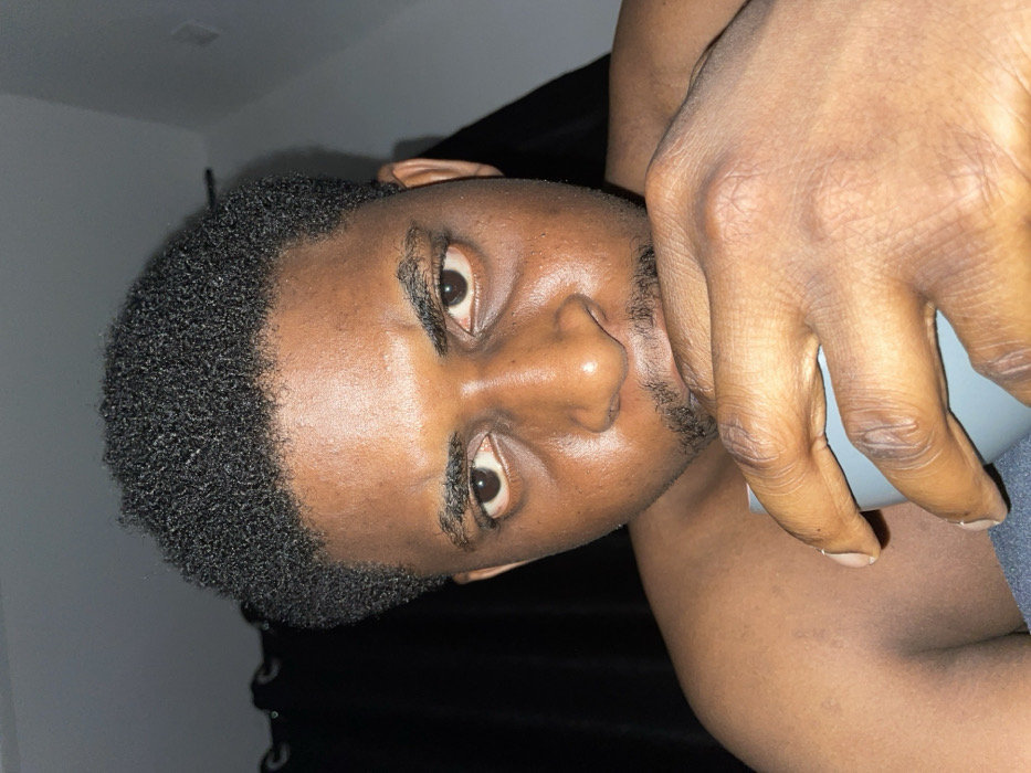 The image displays a close-up, head-and-shoulders view of an adult male in what is identified as the city of Ube, Nigeria. The subject has dark skin, short curly black hair, a thin mustache, and a short beard. His face appears to have an oily sheen, and he exhibits visible under-eye circles. He is shirtless, revealing his bare shoulders. The male holds a light blue or gray mug with his right hand, positioned near his mouth. He stares directly into the camera with an intense expression, his eyebrows slightly furrowed. The background is dimly lit, with a dark curtain visible on the left and a light-colored wall or ceiling section to the upper right.