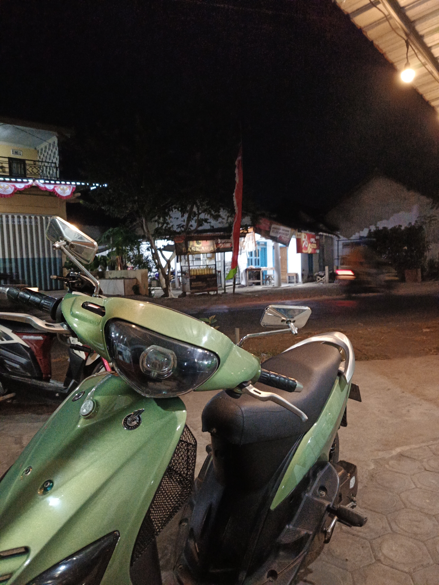 An outdoor, eye-level, medium shot captures a light green scooter parked at night on a paved surface. The scooter is positioned in the foreground, facing right, with its front end prominent. The handle, mirror, and headlight are clearly visible. The background shows a street scene with a building on the left, decorated with white and red banners and lights. Further in the background, there are more buildings with illuminated windows and signs, suggesting a commercial area or shops. A red and white flag is also visible on the left side. The overall lighting suggests evening or nighttime, with a single light bulb hanging from a structure on the right. A blurred light trail on the road indicates vehicle movement. The environment appears to be an urban or semi-urban setting in Sumbersari, Indonesia, possibly a roadside shop or a street market at night. There are no people visible in the scene.