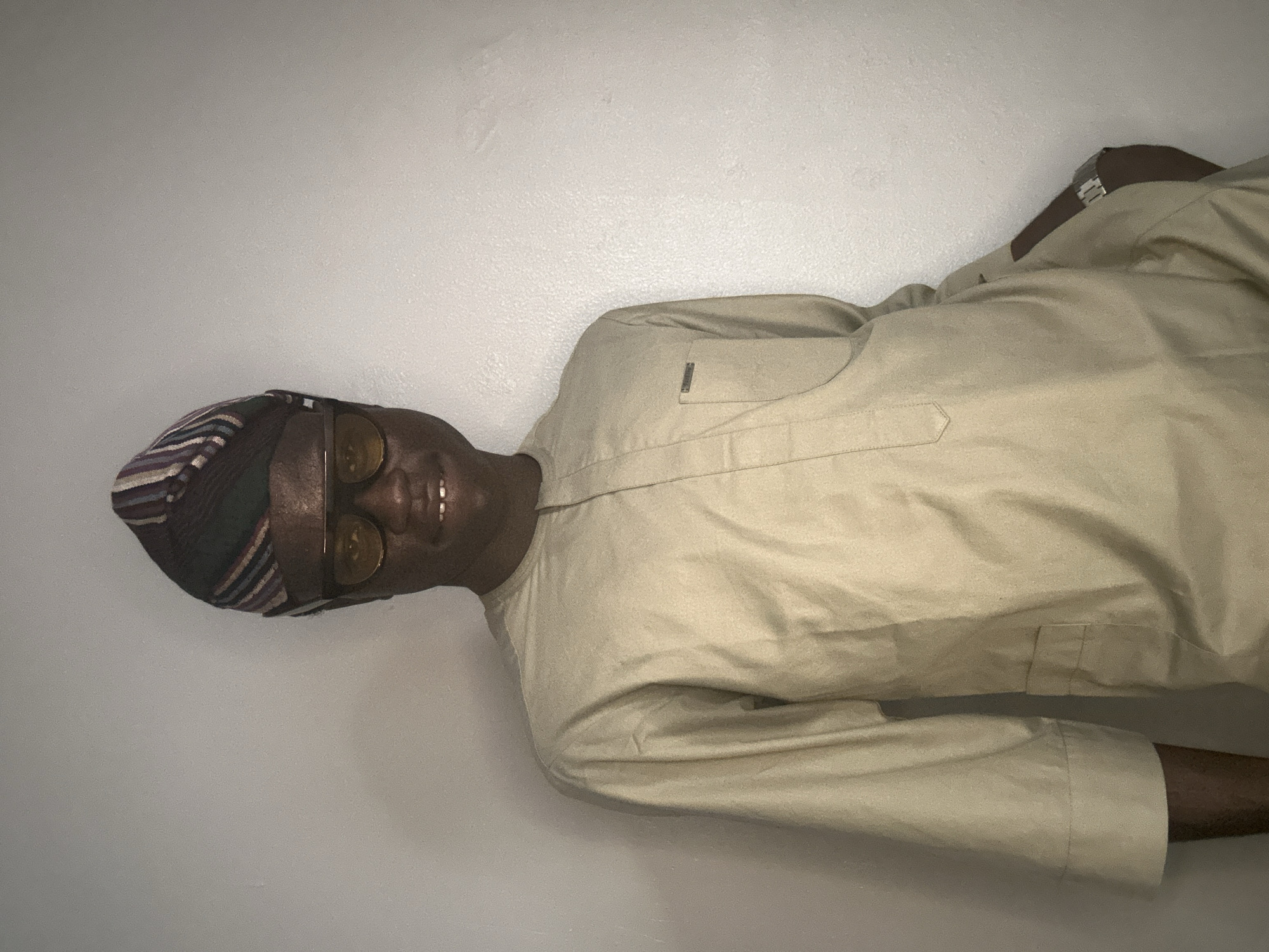 A young Black man is shown from the chest up, standing in front of a plain, lightly textured gray wall. He is wearing a light tan, long-sleeved traditional African garment with a mandarin collar and a single breast pocket on the left side, which has a small metallic tag. On his head, he wears a multi-colored striped Aso Oke cap. He also has on light brown rectangular-shaped sunglasses. He is smiling, showing his teeth. A silver watch is visible on his left wrist. The lighting appears to be coming from the front and slightly from above, casting a subtle shadow on the wall behind him. The image appears to be taken in an indoor setting. The provided context indicates this scene is located in Badagry, Nigeria.