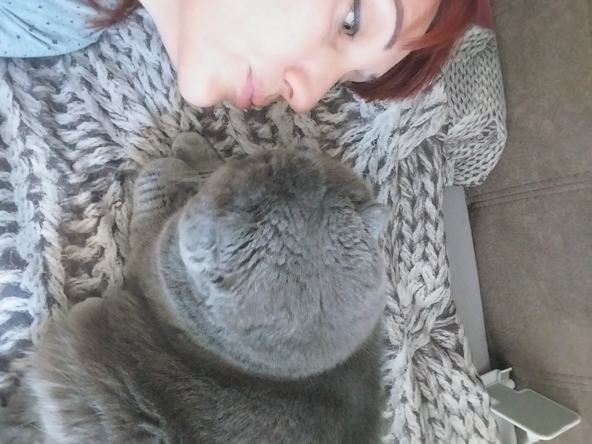 The image shows a close-up, overhead view of a woman and a cat lying together on a couch. The woman is on the left side of the frame, with her face turned towards the cat. She has reddish-brown hair and appears to be about to kiss the cat, with her lips slightly pursed. Her eye is open and looking towards the cat. The cat is on the right side of the frame, facing the woman. It is a large, gray cat with short fur, and its head is turned towards the woman. The cat's body is mostly obscured by its own fur. They are lying on a textured, woven blanket that covers the couch. The couch appears to be a neutral gray color. In the background, a part of a light-colored object, possibly a phone stand, is visible on the couch. The image is taken indoors in what appears to be a domestic setting. There is no information in the image that would suggest a specific location in the city of Dnipro, Ukraine.