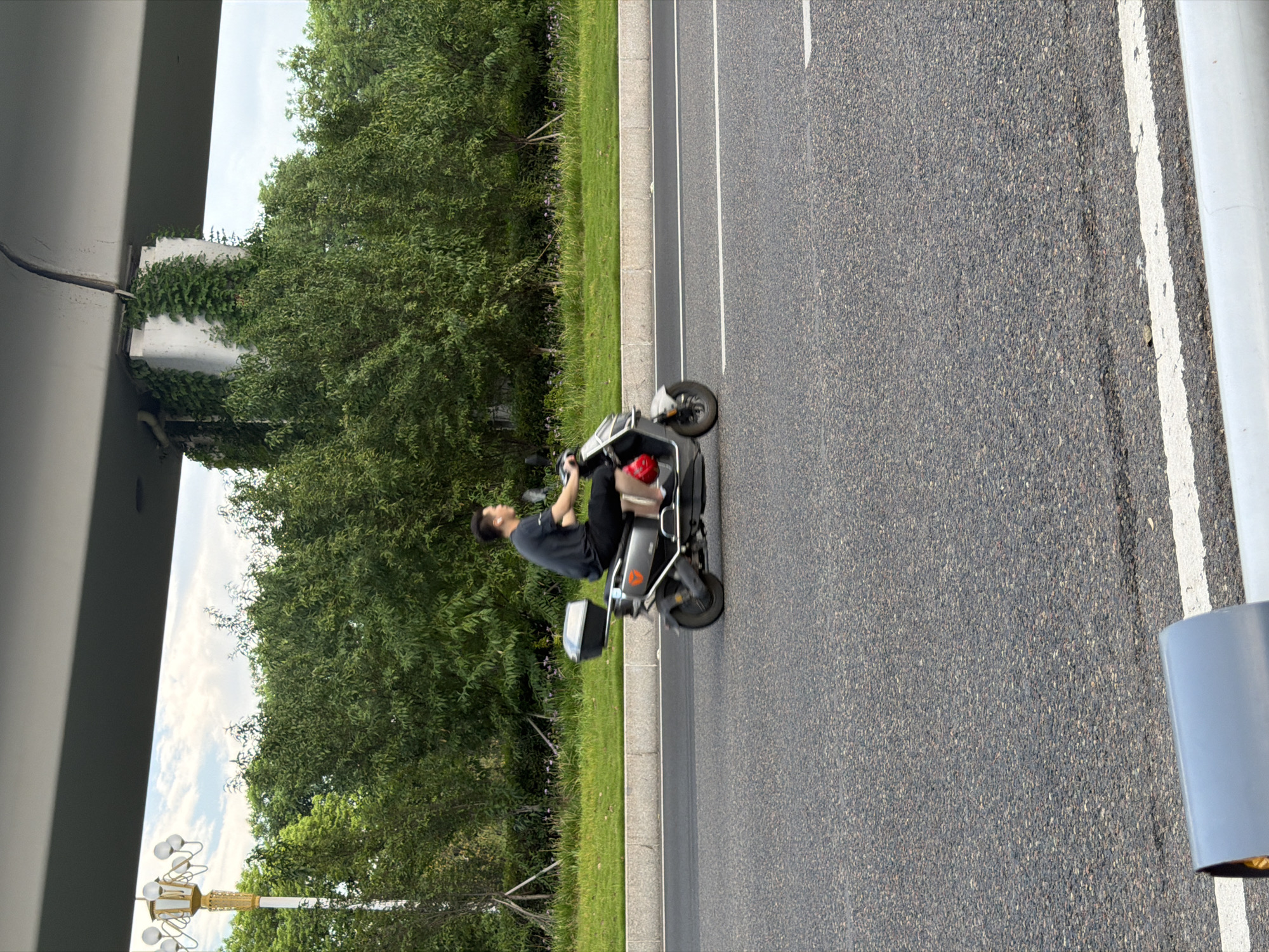 In the city of Jiangjin, China, a person rides an electric scooter on an asphalt road. The scooter is grey and has a black storage box mounted on the back. The rider is wearing a dark t-shirt and shorts, and has headphones in their ears. In the background, there is a line of green trees, a lamppost, and a concrete structure covered in vines, which appears to be a part of an overpass. The sky is cloudy with some patches of blue. A white line is painted on the road, indicating a lane marker. The image captures a moment in time on a city street.