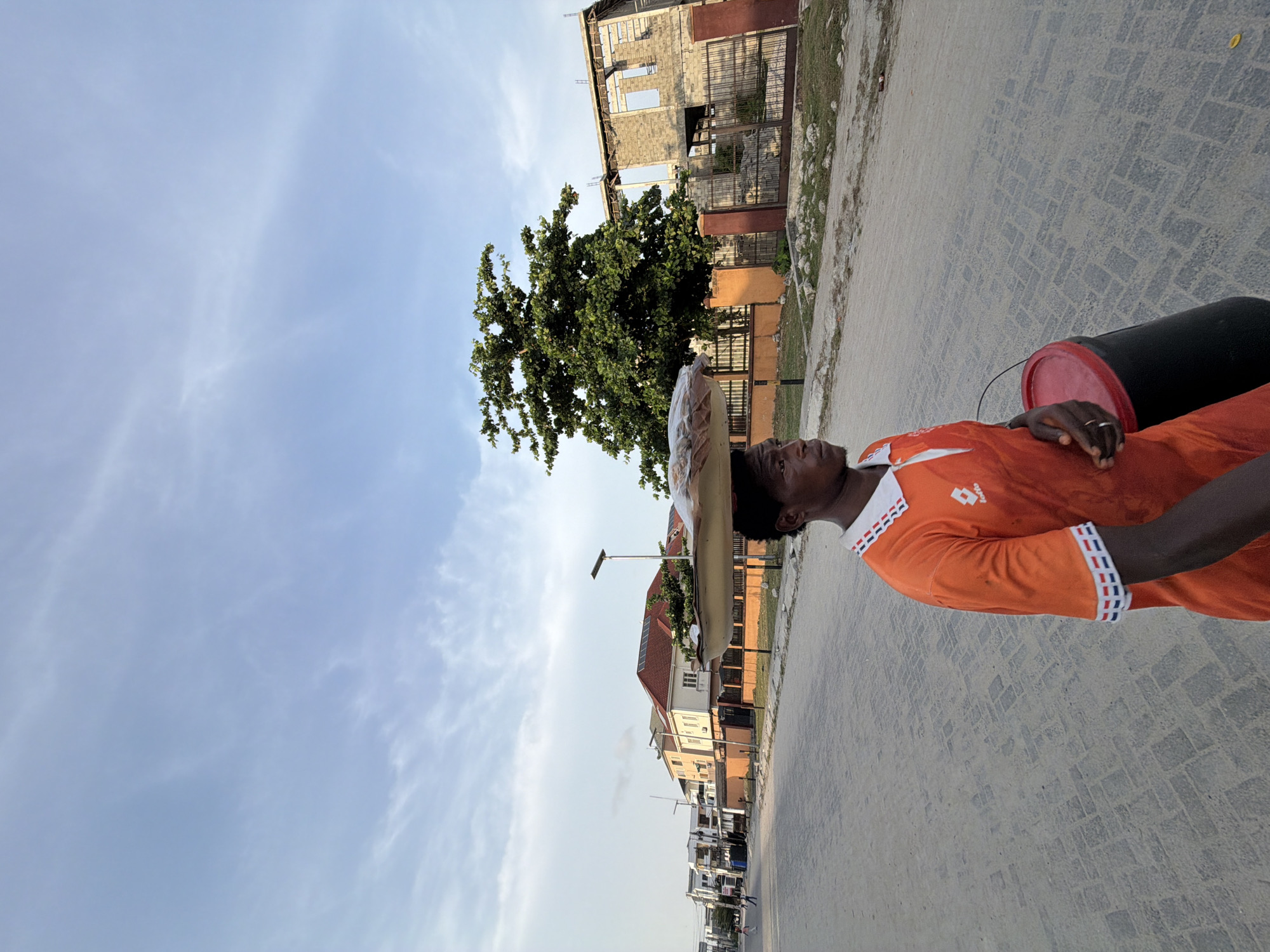 In Igboefon, Nigeria, a man walks along a cobblestone street with a large container and a wrapped item balanced on his head. He is wearing an orange shirt with white and red accents. Behind him are buildings in varying states of construction and a row of houses along the street. The sky is partly cloudy.