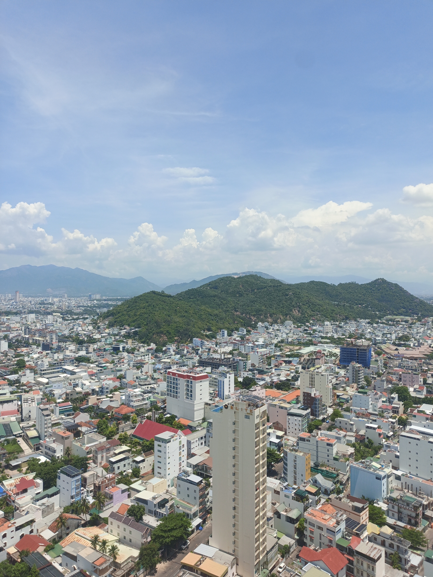 This is an aerial, high-angle photograph capturing a dense urban landscape with lush green mountains in the background under a bright, partly cloudy sky. The city below is densely populated with a mix of low-rise and mid-rise buildings, predominantly white and light-colored, with scattered red-tiled roofs. Numerous trees are visible interspersed between the buildings, indicating a tropical or subtropical climate. In the foreground, a tall, slender, light-colored building stands out amongst the lower structures. The image appears to be taken during daylight hours, with the sun casting subtle shadows. The scene depicts a typical view of Bac Nha Trang Ward, Vietnam, showcasing its coastal proximity and mountainous terrain meeting the urban sprawl. There are no visible people or specific actions depicted in the image.