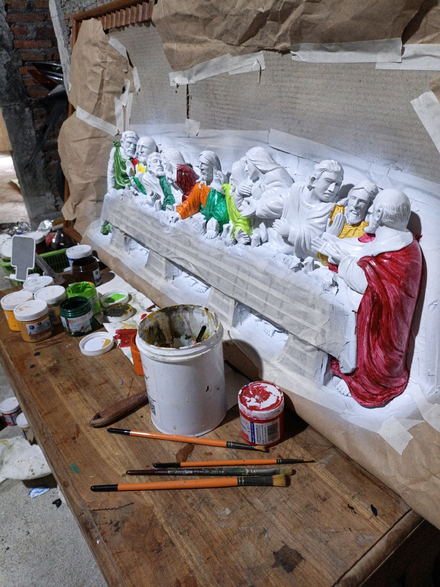 A statue of the Last Supper is being painted on a wooden table in an outdoor workshop in Cikal, Indonesia. The statue is mostly white, with some areas already painted in red, green, and yellow. Several paint cans and brushes are scattered on the table around the statue. The background shows a brick wall and brown paper.