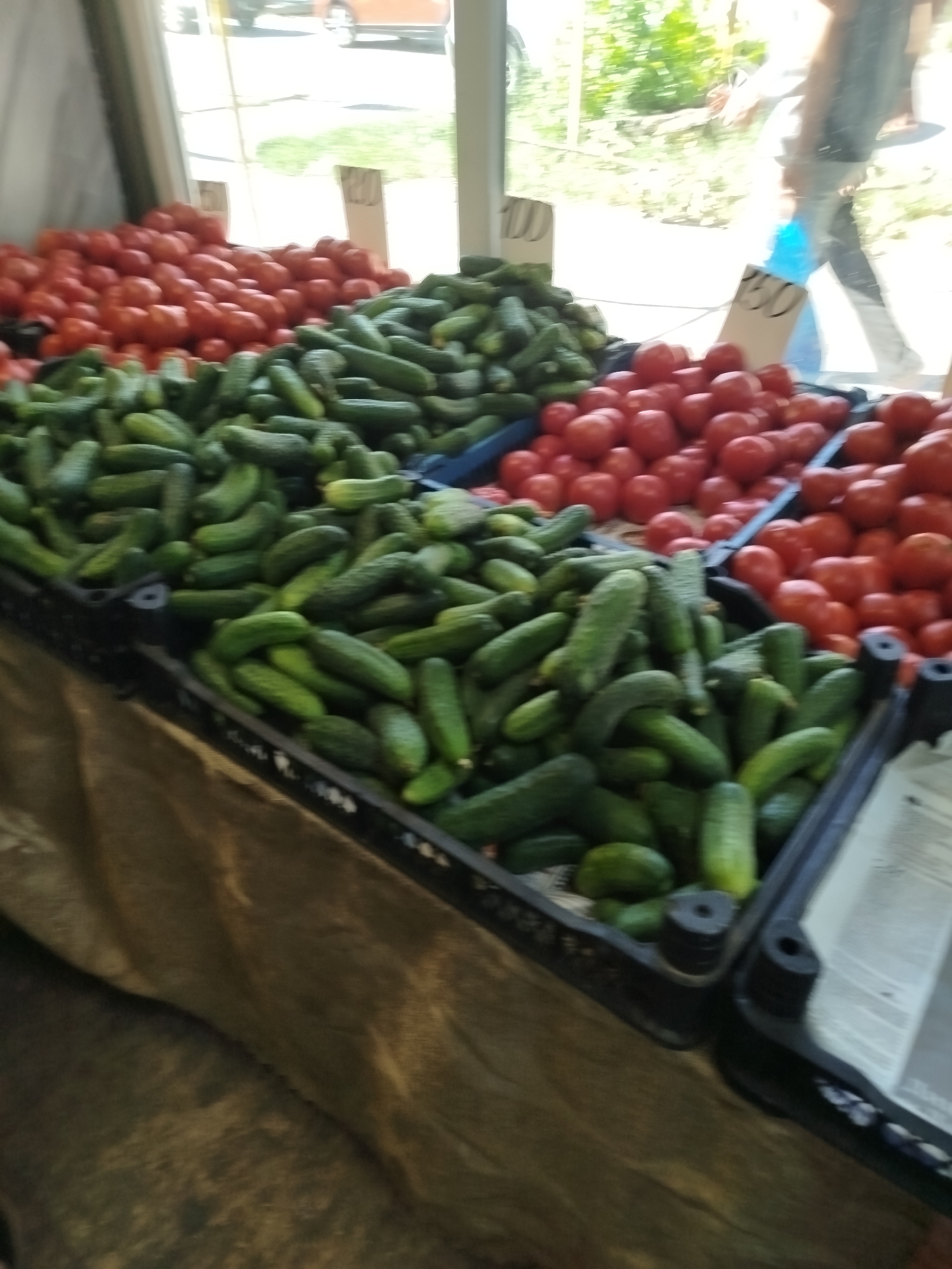 The image depicts a market stall selling fresh produce, specifically a large quantity of tomatoes and cucumbers. The stall is located in Taganrog, Russia, as evidenced by a sign in the background with Russian writing. The setting appears to be outdoors, possibly a street market or a farmer's market, with a window or opening to the right showing greenery and a person walking in the distance. The produce is arranged in large bins, with tomatoes in the background and cucumbers in the foreground. There are also price tags visible, one with "100" written on it. The overall impression is one of a vibrant, bustling market scene.