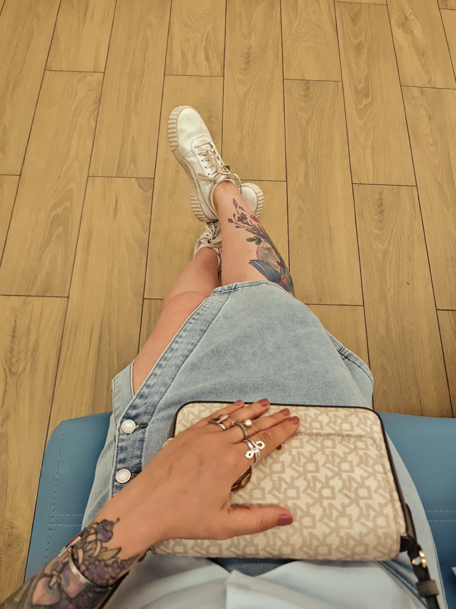 A high-angle, first-person perspective photograph captures a person sitting in an indoor setting. The individual is seen from the waist down, wearing a light blue denim pinafore dress or overall skirt with "TOMMY JEANS" visible on the silver buttons. Their legs are crossed at the ankles, showcasing white lace-up sneakers with thick, off-white soles.
The person has multiple tattoos: a large, colorful design of flowers and possibly a bird or moon on the left thigh, and a smaller, geometric design on the right ankle. A sleeve tattoo with purple and green floral motifs is visible on the left forearm.
The right hand rests on a light beige or cream-colored handbag with a repetitive "DKNY" monogram pattern. The hand is adorned with several silver rings, including one with a pearl and another with a red, white, and blue stripe. The fingernails are painted a dark pink or mauve.
The person is seated on a light blue upholstered chair, part of which is visible at the bottom and sides of the frame. The background consists of a clean, light-colored wooden floor with rectangular planks laid in a staggered pattern. The activity depicts a moment of rest or waiting. The location context provided is Moscow, Russia.