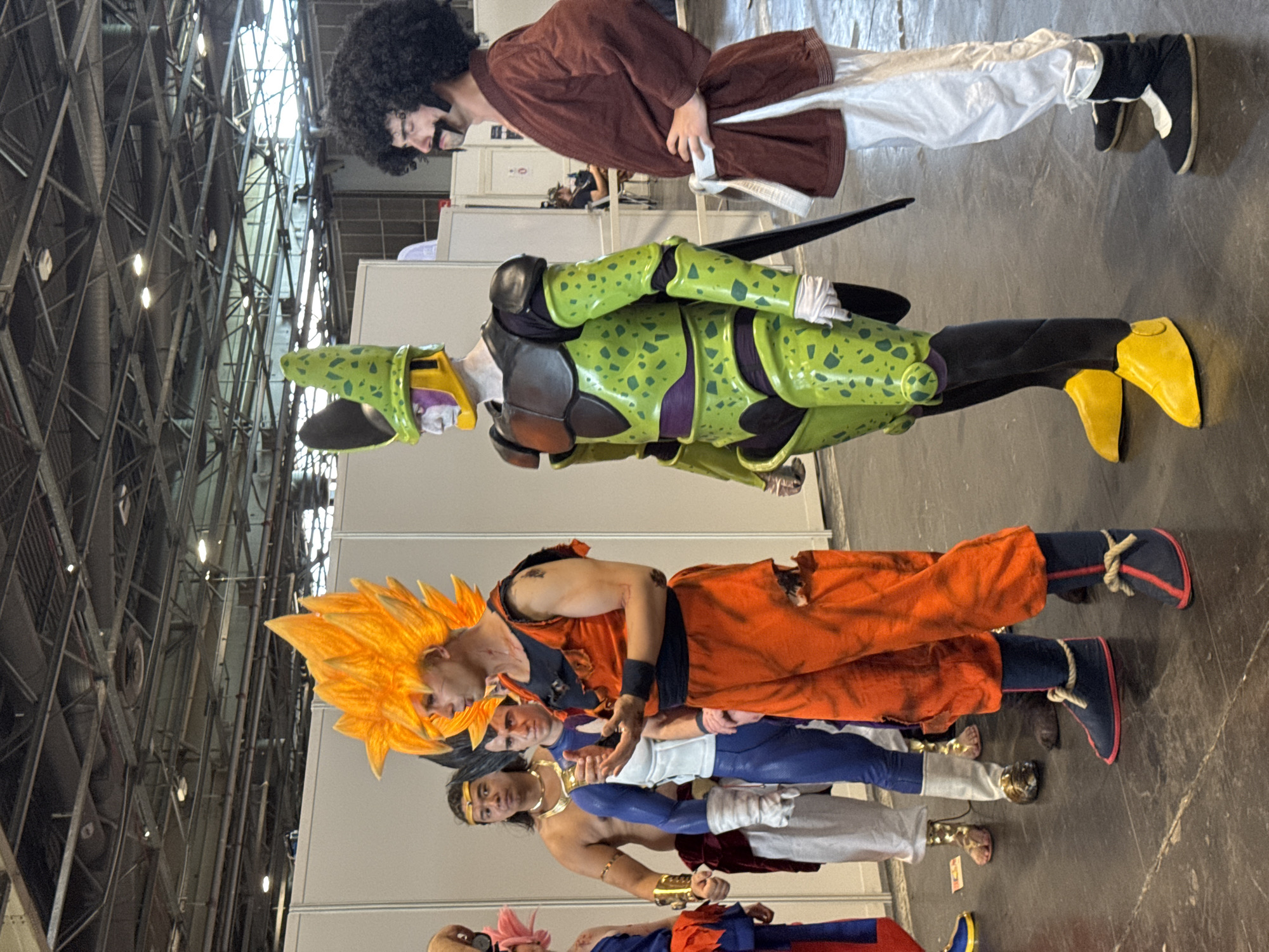 The image depicts a group of people in cosplay at an indoor event, likely a convention, in Villepinte, France. The subjects are standing in a line on a dark grey floor. The person in the foreground to the right is dressed as Goku from Dragon Ball Z, wearing an orange gi with ripped sections and a spiky orange wig. To their left is a person in a green and black costume with a distinctive helmet, resembling Cell from Dragon Ball Z. Further to the left are individuals dressed as other Dragon Ball Z characters, including one with a muscular physique and a golden necklace, and another in a blue and white suit. On the far right, a person with a large afro wig and a mustache is dressed in a brown top and white pants. The background consists of white partition walls and the overhead structure of a large hall with exposed beams and lighting.