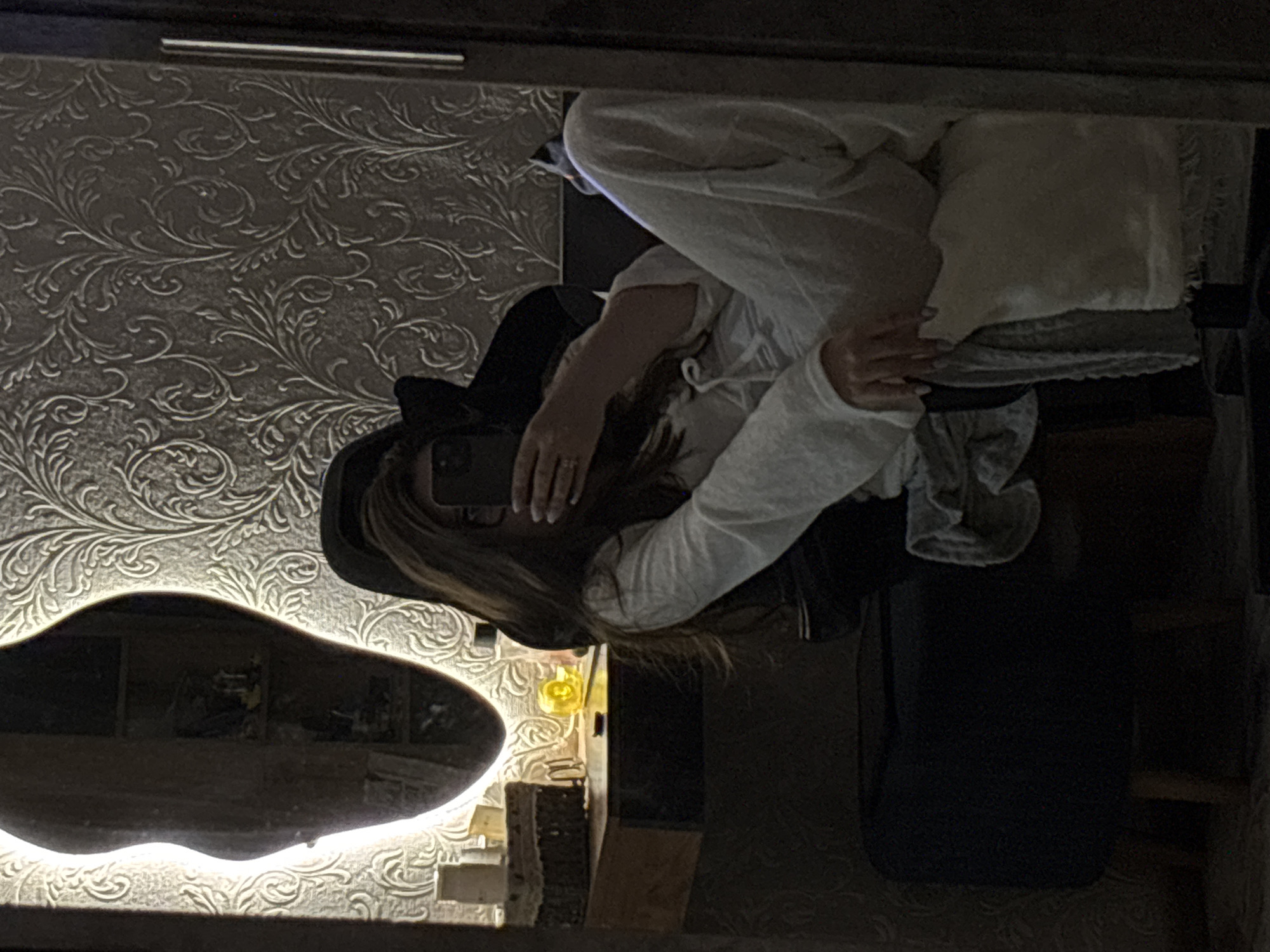 A reflection in a mirror shows a person wearing a black hat and white loungewear. The individual is positioned on a surface covered with a white pillow and gray bedding. The background features a wall with an ornate, floral pattern. In the lower-left corner of the mirror's reflection, there is a blurred image of an interior scene, including a glowing light fixture and other objects. The setting appears to be indoors, possibly a bedroom. It is not possible to determine the specific location in Chaikovsky, Russia, from the image alone.