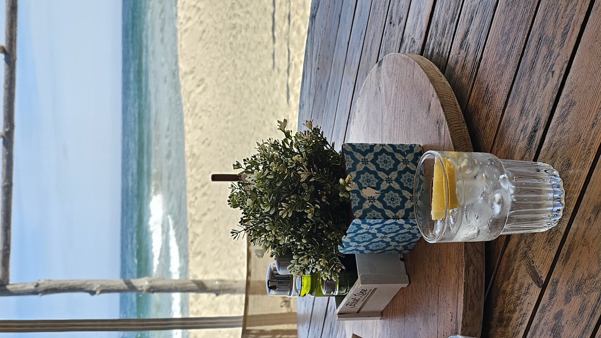 The image captures a serene seaside setting from what appears to be a restaurant or cafe in Kranevo, Bulgaria, looking out over the Black Sea. The photo is oriented vertically, showing a table surface on the right and the ocean view through a window or opening on the left.
In the foreground, a rustic wooden table with distinct grain lines occupies the right side of the frame. On the table, a clear glass filled with ice and a lemon wedge suggests a refreshing drink. To its left, a small, vibrant green artificial plant is housed in a decorative pot featuring blue and white geometric patterns. Next to the plant, there's a small wooden caddy containing what appear to be salt and pepper shakers or oil and vinegar bottles. The word "First Line" is visible in white script on the front of this wooden caddy. A round wooden cutting board or decorative piece is partially visible behind the plant.
Through the window or opening on the left, a bright, sunny daytime scene unfolds. The tranquil blue waters of the sea meet a light sandy beach, with gentle white waves breaking near the shore. A wooden beam or frame, likely part of the establishment's structure, runs vertically along the far left edge of the image. The overall impression is one of relaxation and enjoying a meal or drink with a beautiful ocean view.