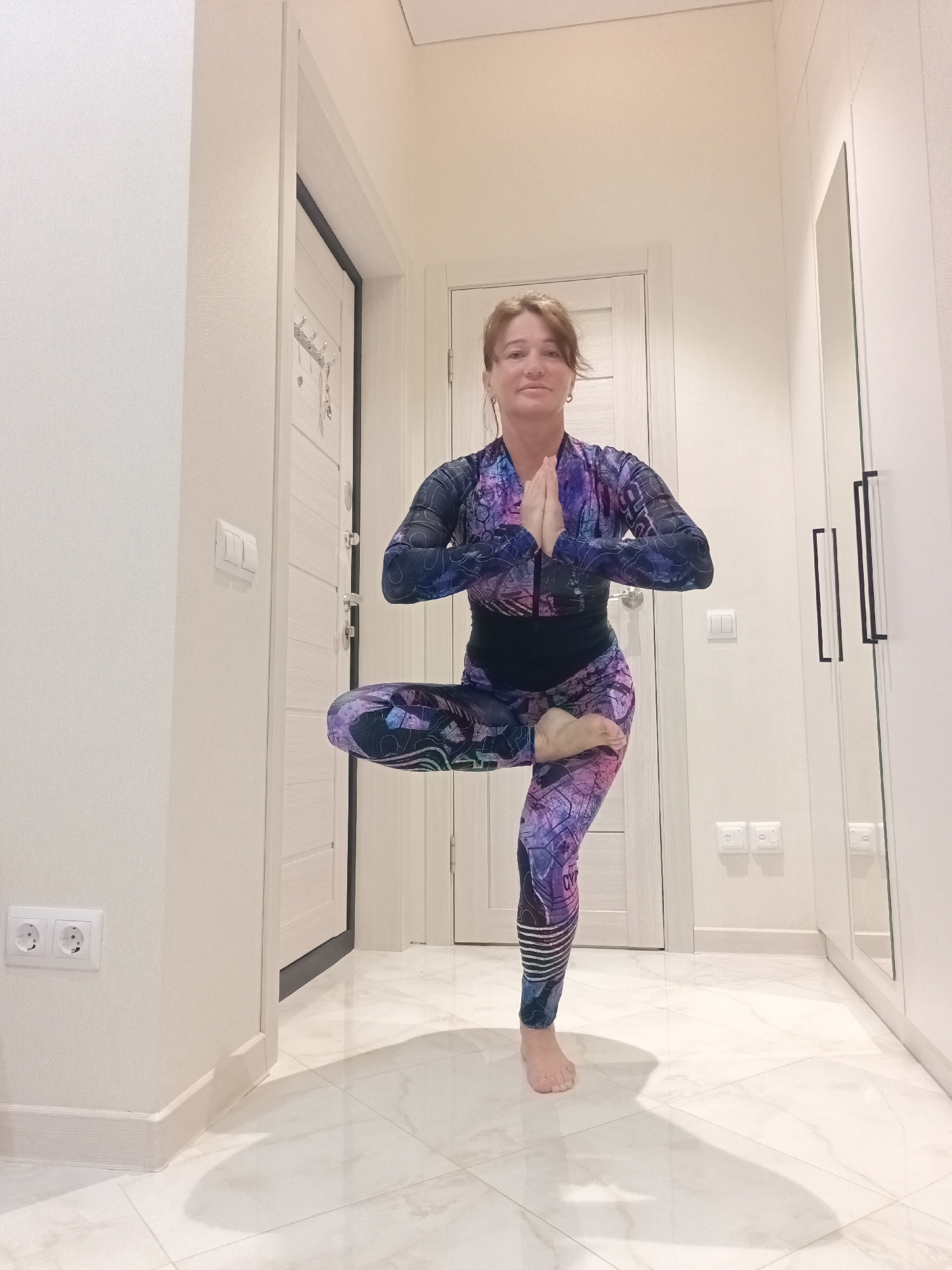 A woman is photographed indoors in a hallway, performing a yoga pose. She is standing on her right leg with her left foot placed on her right thigh, in what appears to be a variation of the Tree Pose (Vrksasana). Her hands are pressed together in front of her chest in Anjali Mudra. She is wearing a long-sleeved, full-length yoga suit with a vibrant purple and blue abstract pattern. The hallway features cream-colored walls, white doors, and a light-colored tiled floor with a reflective sheen. A large white wardrobe with black handles is visible on the right side of the frame, and a full-length mirror reflects a portion of the room. Electrical outlets are visible on the walls. The lighting in the hallway is soft and diffused. The scene does not contain any specific indicators of location within Sochi, Russia.
