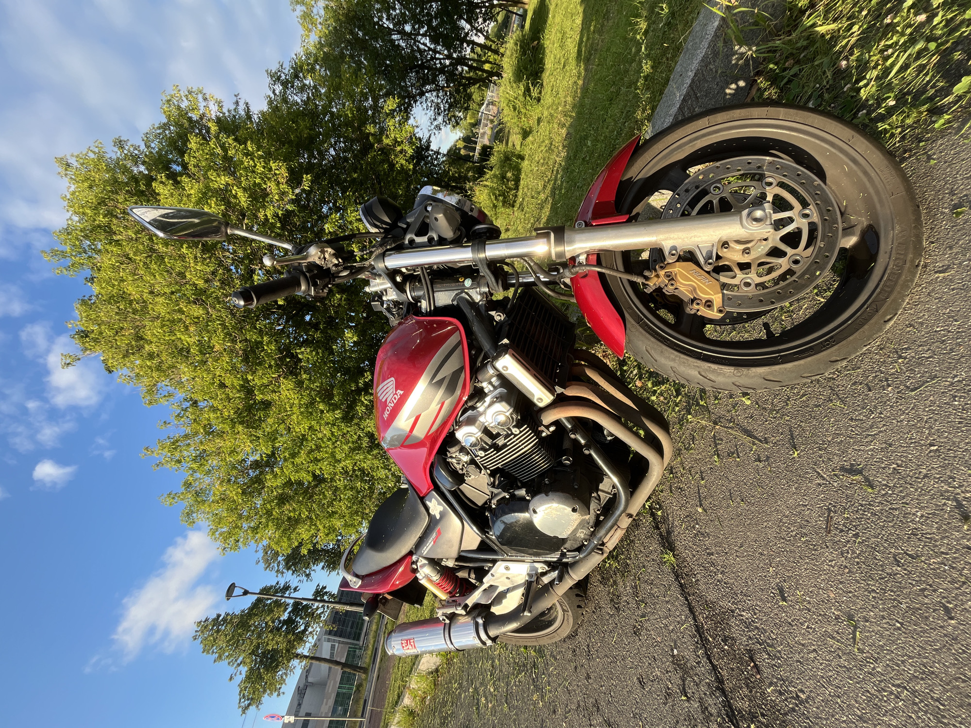A red Honda motorcycle is parked on an asphalt surface next to a grassy area with a large tree. The motorcycle is a street bike with a visible engine, exhaust pipes, and a shiny chrome handlebar. The front wheel and part of the brake system are prominently featured. In the background, there are more trees, a bright blue sky with scattered white clouds, and a distant building. A lamppost is also visible. The scene appears to be set during the daytime, likely late afternoon due to the low angle of the sun casting long shadows. The weather seems clear and pleasant. The image has a slightly tilted perspective. Visible text includes "HONDA" on the fuel tank and "YOSHIMURA" on the exhaust. The overall mood is relaxed, suggesting the motorcycle is casually parked outdoors.