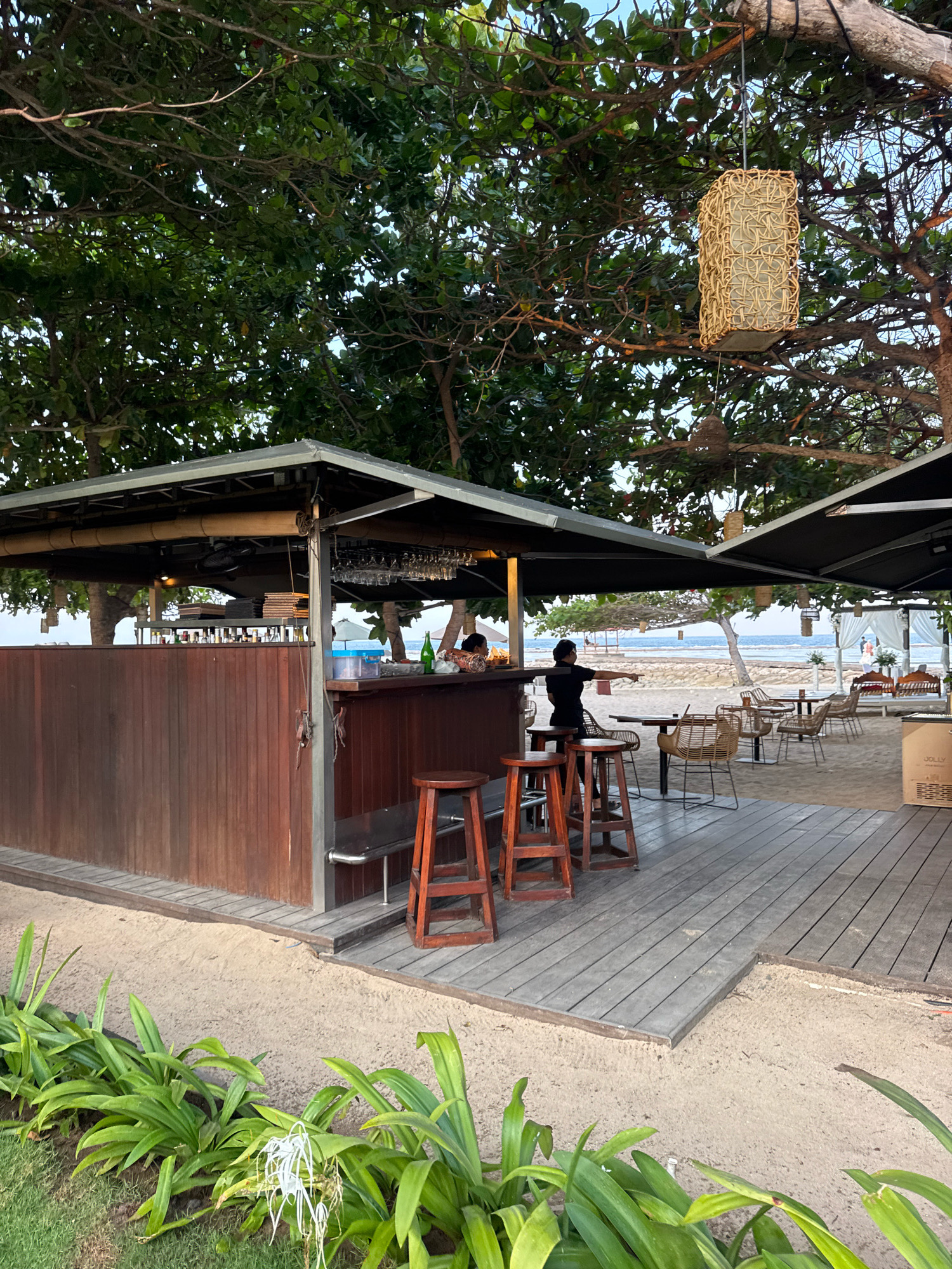 This image depicts an outdoor beachfront bar and dining area in Kuta Selatan, Indonesia. A wooden bar with stools is set up on a sandy surface, partially covered by a dark awning. Trees with dense green foliage overhang the area, with a woven lamp hanging from a branch. In the background, several tables and chairs are arranged on a wooden deck, leading towards a beach and the ocean. A person is seen standing behind the bar, and another is seated at a table. The environment appears to be a resort or a casual eatery, with lush vegetation in the foreground.