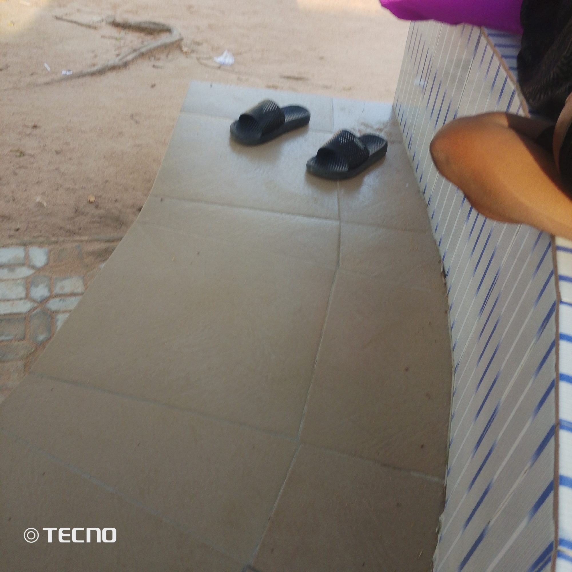 The image depicts an outdoor scene in Jalingo, Nigeria. In the foreground, a tiled surface, likely part of a bench or walkway, is visible. Two black slide sandals are placed on this tiled surface. The background consists of a sandy area with some debris. To the right of the tiled surface, a person's leg and thigh, exhibiting tanned skin, are partially visible, suggesting they are sitting or resting. The person is wearing dark clothing. A portion of a wall with a striped pattern of blue and white is also visible, forming a boundary for the tiled area. The "TECNO" logo is present in the bottom left corner of the image.