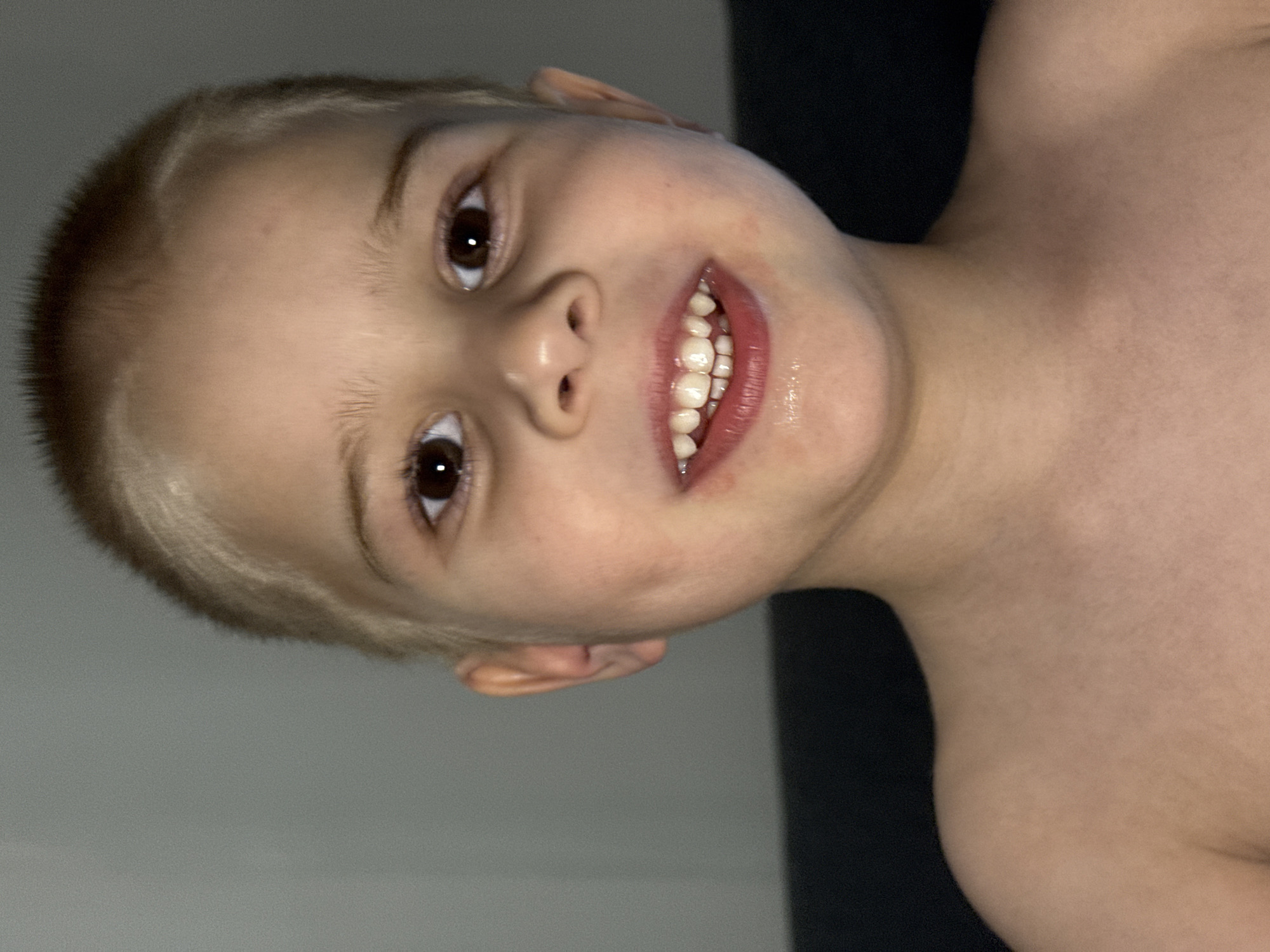 A close-up portrait shows a young boy with a fair complexion, short blonde hair, and brown eyes. He is smiling, revealing his teeth. The background is divided between a gray wall on the left and a dark, possibly black, surface on the right. The boy's upper chest and shoulder are visible. The location is not identifiable from the image. The city of Chaikovsky, Russia cannot be inferred from the content of the image.