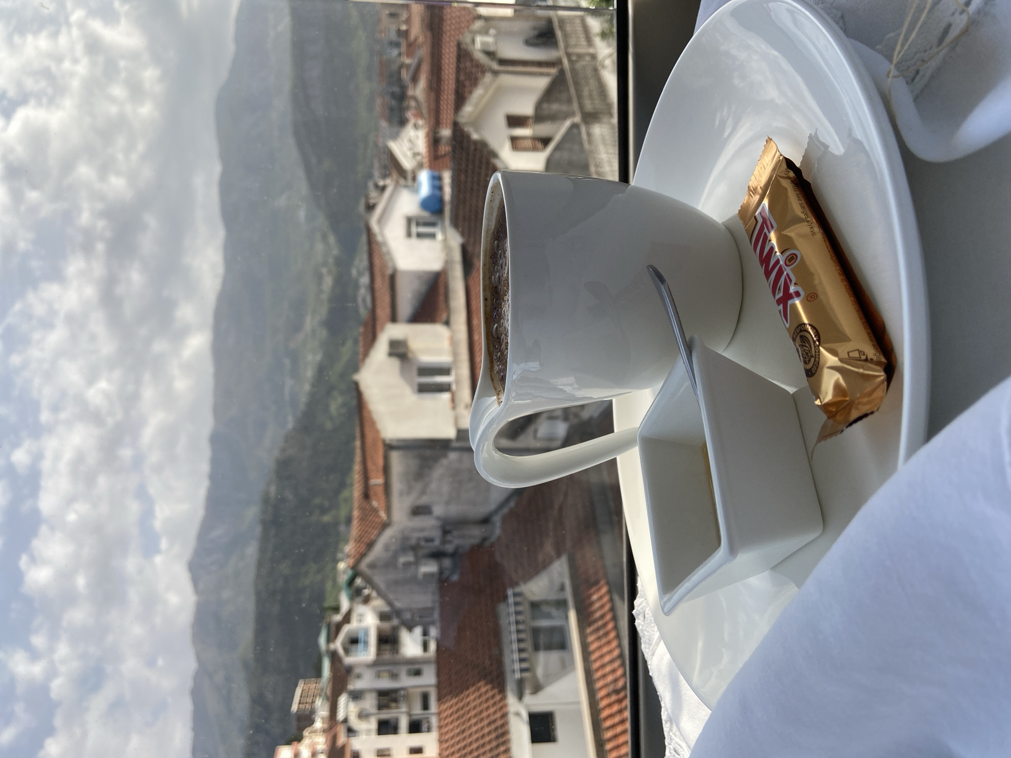 A bird's-eye view reveals a coffee cup, condiment holder, and a wrapped Twix candy bar arranged on a white plate. A white cloth partially obscures the right side of the scene. Outside the window, the cityscape of Budva, Montenegro, unfolds against a backdrop of distant mountains and a cloudy sky. Buildings with red-tiled roofs and white facades are discernible, typical of the region's architecture.