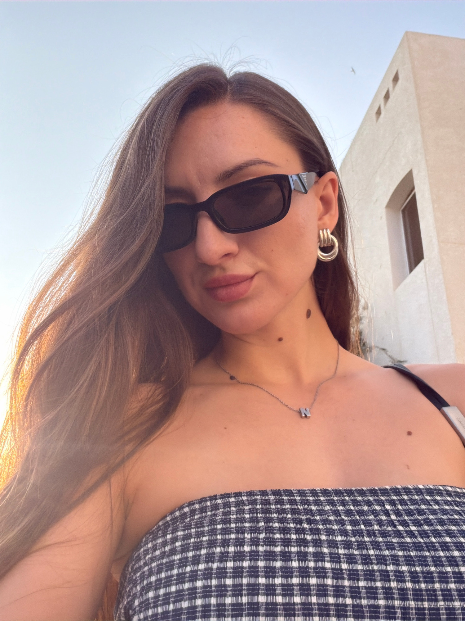 A woman wearing sunglasses, a strapless checkered top, and a necklace with the letter 'N' is posing for a selfie. She has long brown hair that is partially obscuring her face and shoulder. A building with white walls and windows is visible in the background, along with a clear sky. The image is taken outdoors, possibly in the city of Sharm El Sheikh, Egypt, during the daytime, as indicated by the bright sunlight.