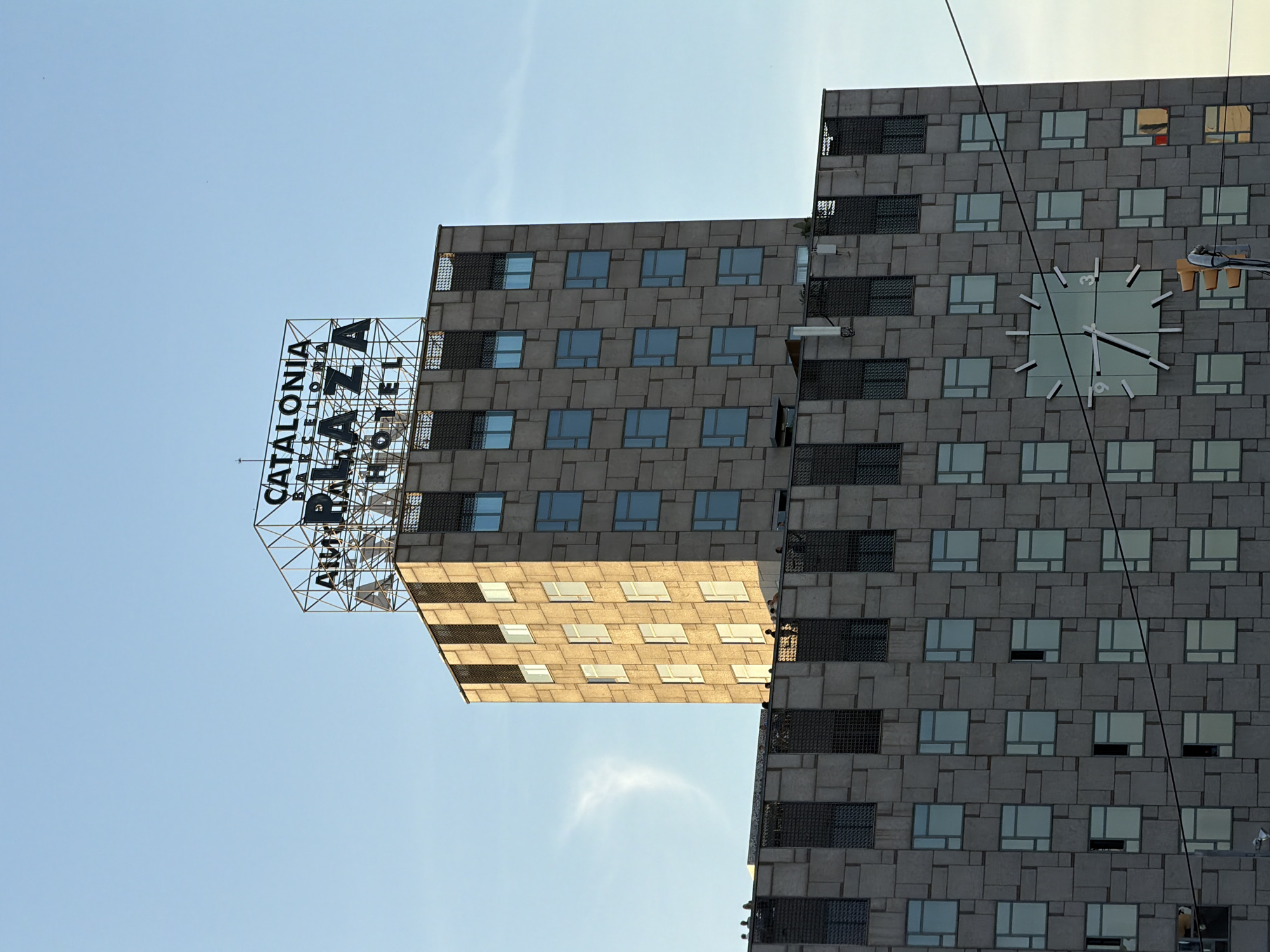 The image shows the Catalonia Plaza Hotel in Barcelona, Spain, under a clear blue sky. The hotel has a distinctive Brutalist architectural style, with a facade composed of grey stone tiles of varying sizes. The building is characterized by a repetitive pattern of windows, many of which are tinted blue, and vertical grates. A prominent feature is a large, modern clock face integrated into the building's facade, with minimalist white hands and markers. The hotel's name, "CATALONIA PLAZA HOTEL," is displayed in large, dark lettering on a metal frame atop the building, with "BARCELONA" written in smaller letters above "PLAZA." In the foreground, a traffic light and a portion of a power line are visible. A few people are seen on a rooftop terrace, silhouetted against the sky. The overall scene captures a segment of the urban landscape of Barcelona.