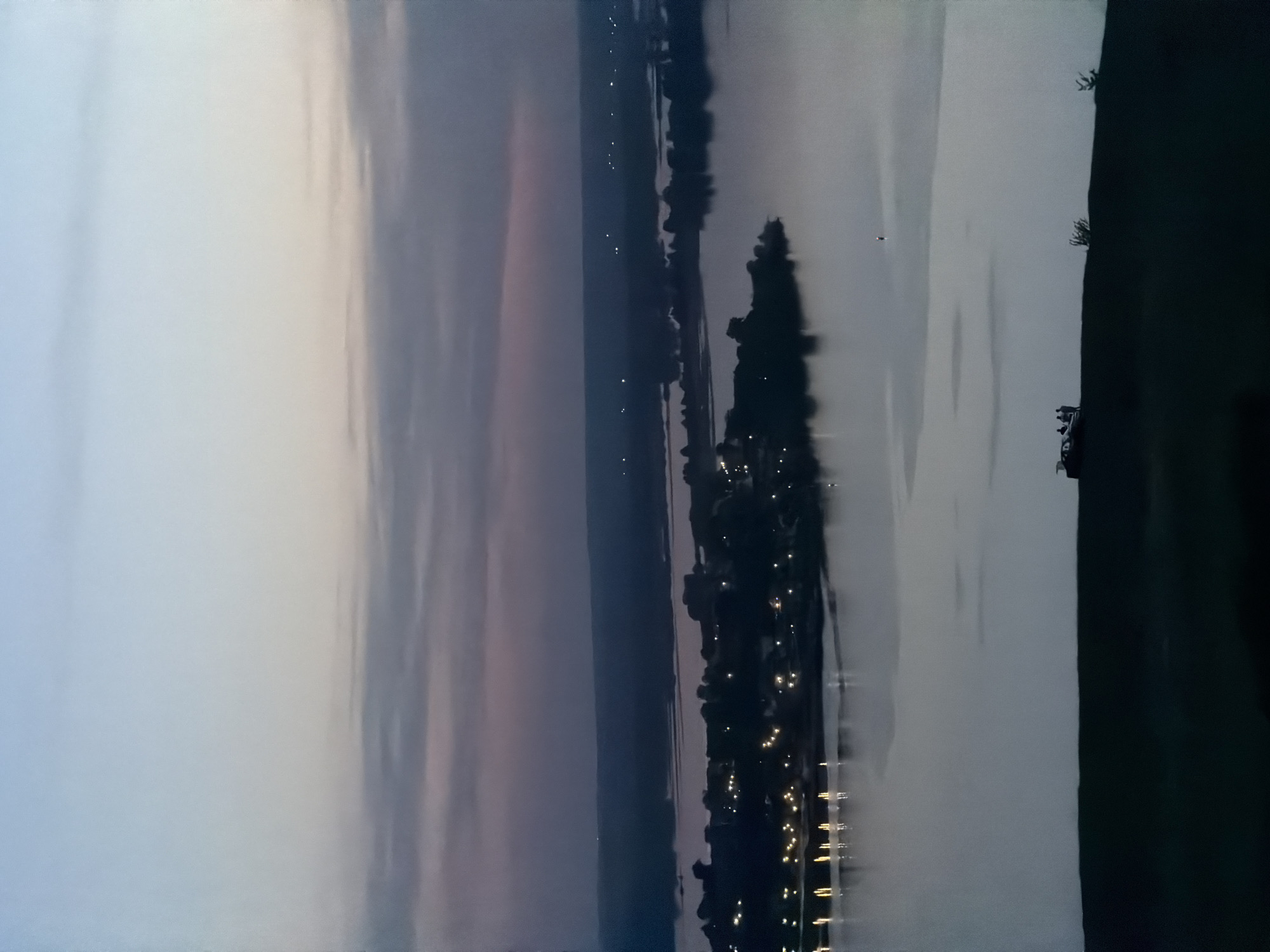 The image depicts a waterscape in Innopolis, Russia, likely at dusk or dawn. A body of water, possibly a river or lake, dominates the scene with reflected lights indicating a settlement along its far bank. The sky exhibits varied tones, suggesting twilight conditions. Land is visible on the right side, with dark foliage suggesting trees or bushes. Lights are visible on the opposite bank, possibly streetlights or building lights, indicating a town or city. The overall mood is serene.