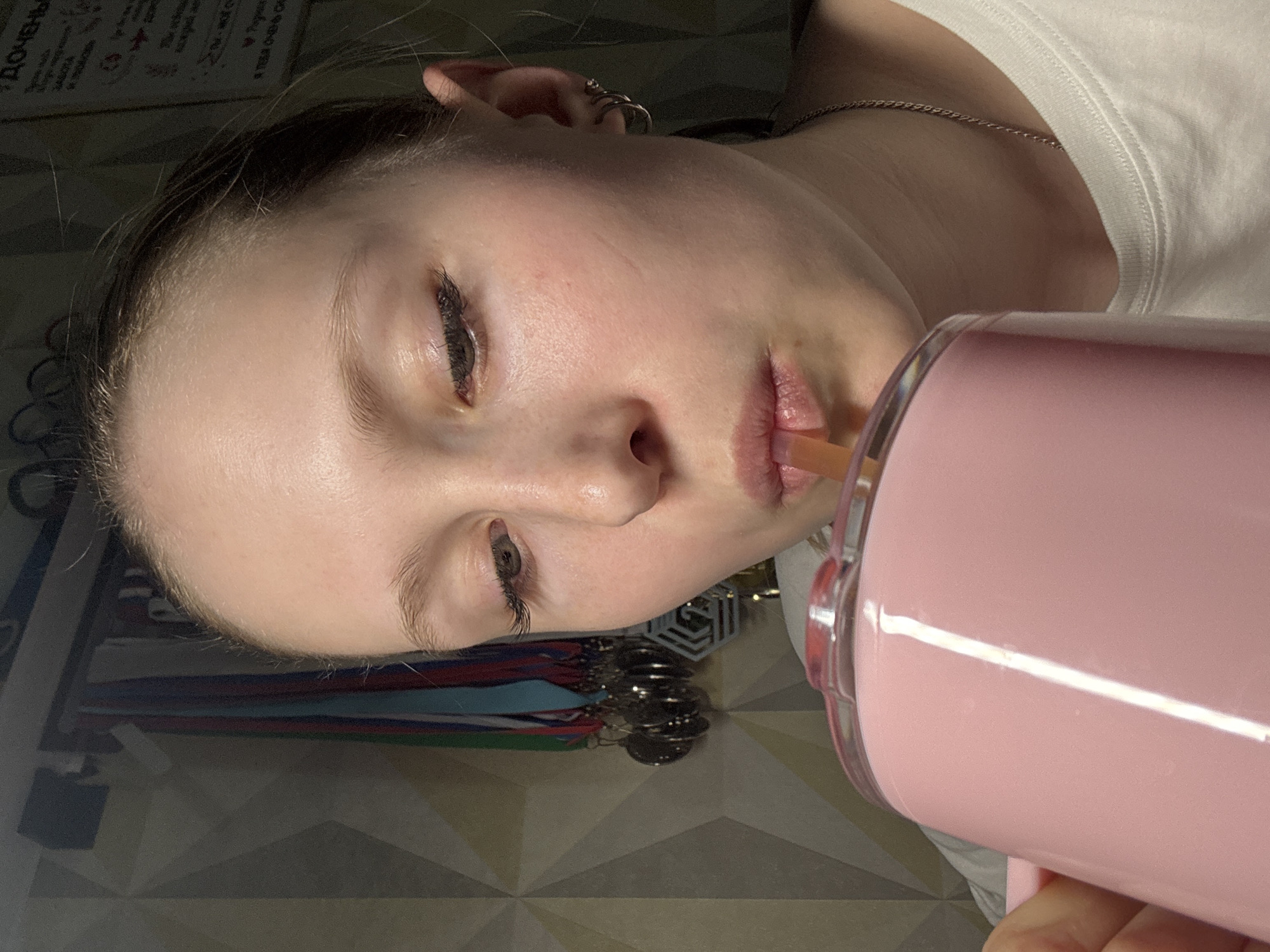 A person is pictured indoors, drinking from a pink cup with a straw. The person has fair skin, closed eyes, and light eyebrows and eyelashes. They are wearing a white t-shirt and a silver necklace. The background includes a wall with items hanging on it, and a floor with a patterned design. The location is possibly Chaikovsky, Russia.